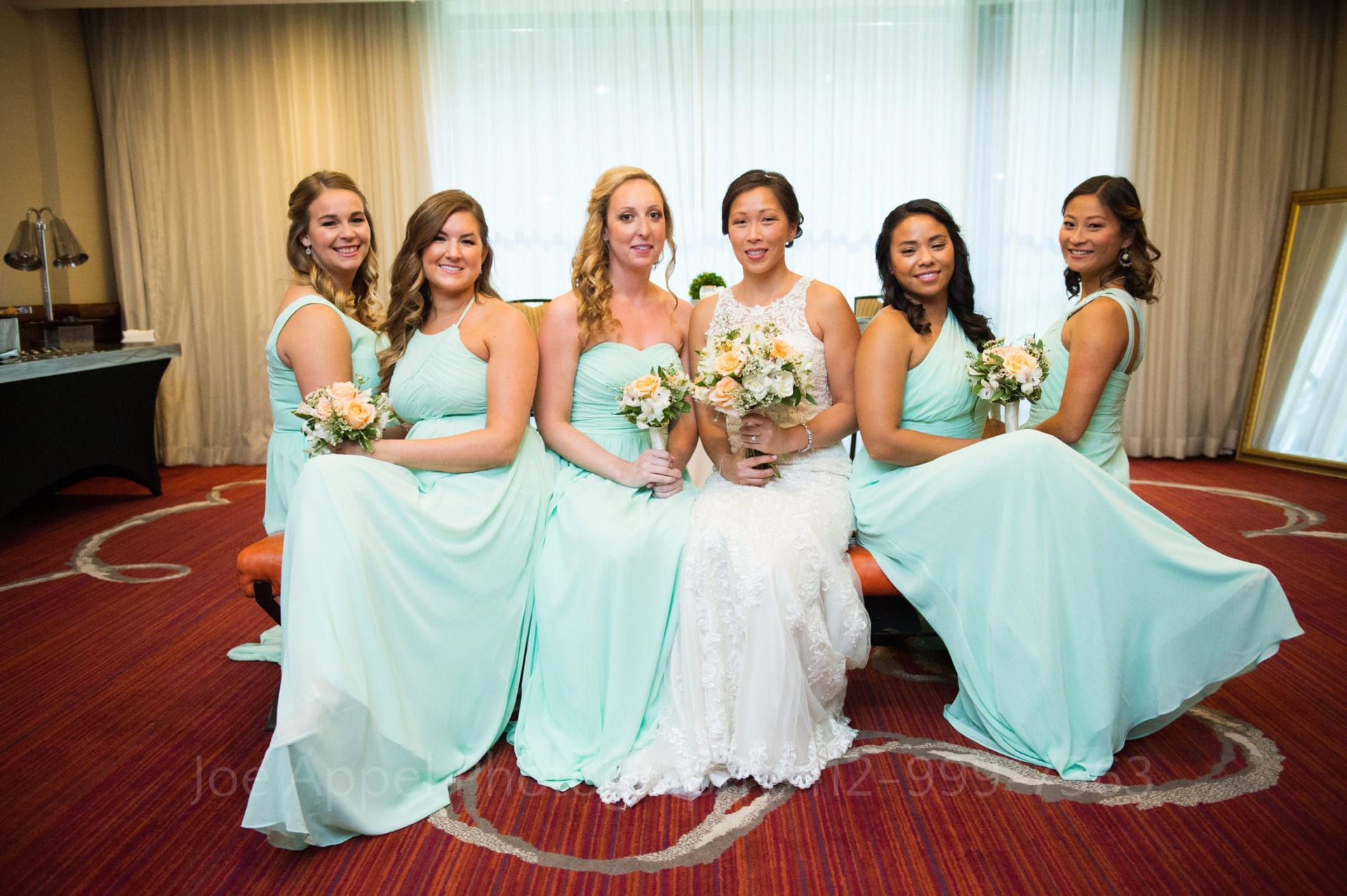 bridesmaids wearing aqua dresses and holding orange bouquets sit on a couch with the bride