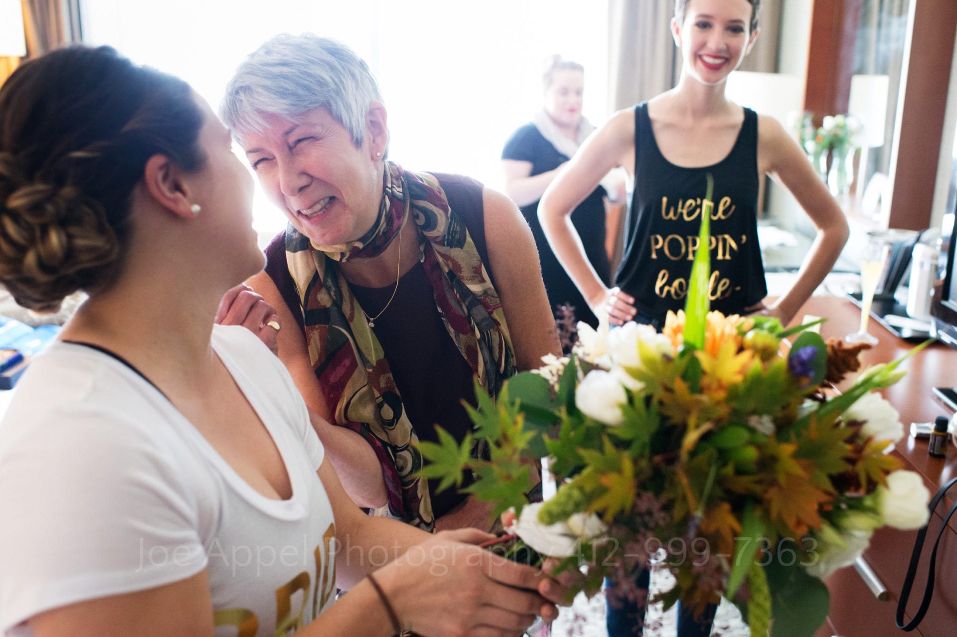 A woman holds a bouquet of flowers as an older woman leans over to her and smiles. Another woman in a black tank top stands at the rear of the frame.