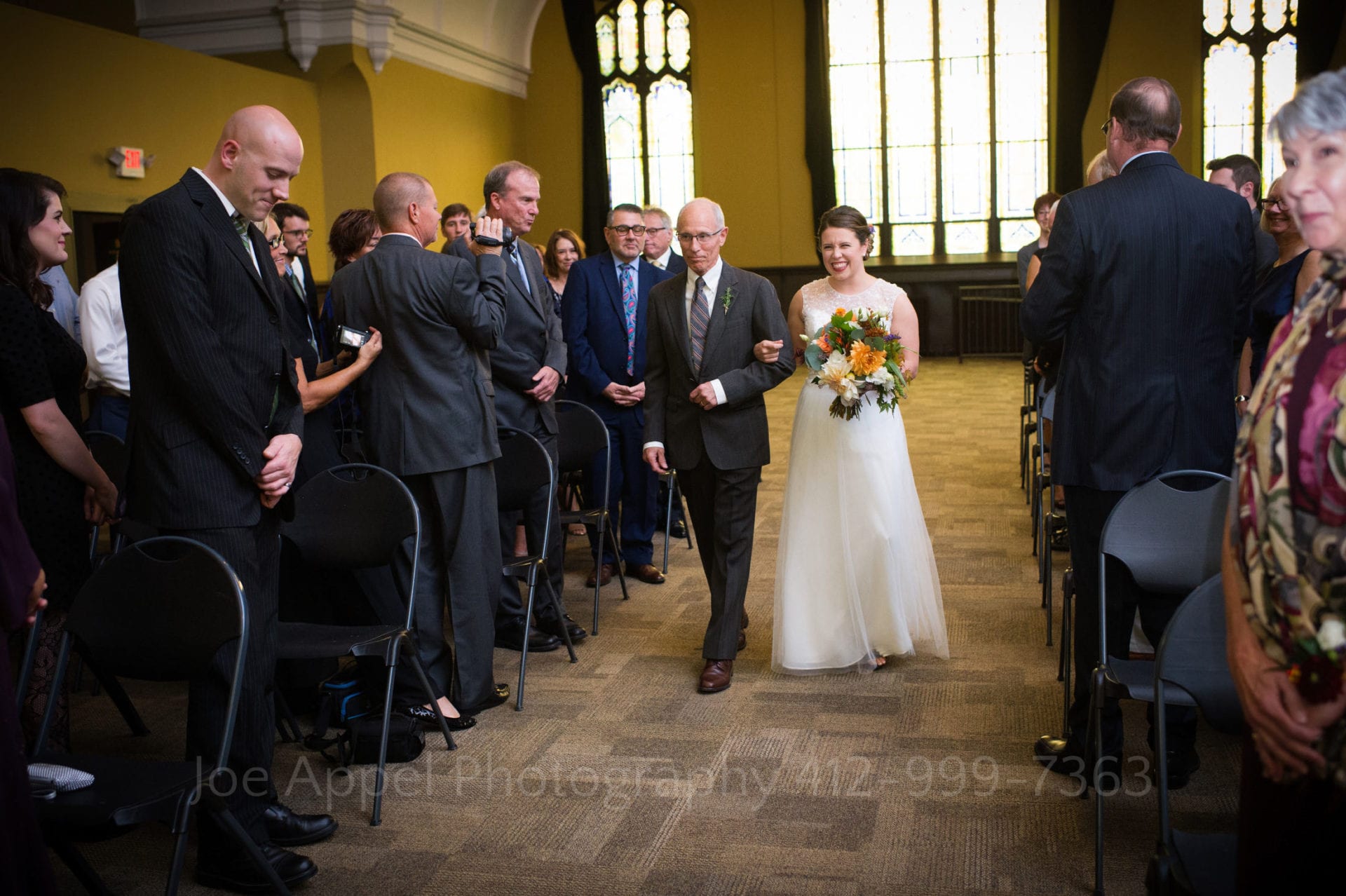 A bride walks down the aisle with her father as guests stand on either side of them. The walls in the room are mustard colored.