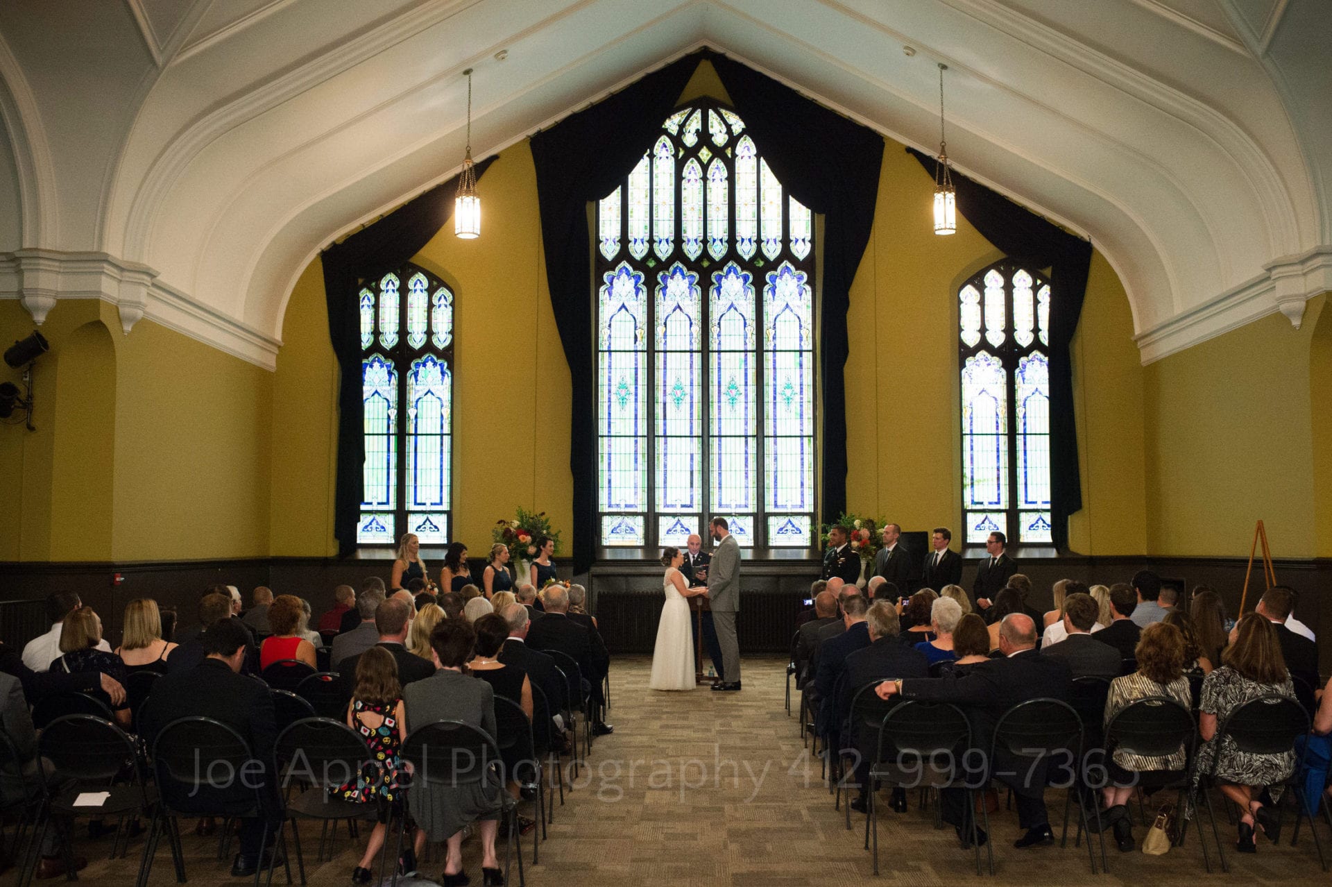A bride and groom stand at the front of their ceremony with large stained glass windows behind them.