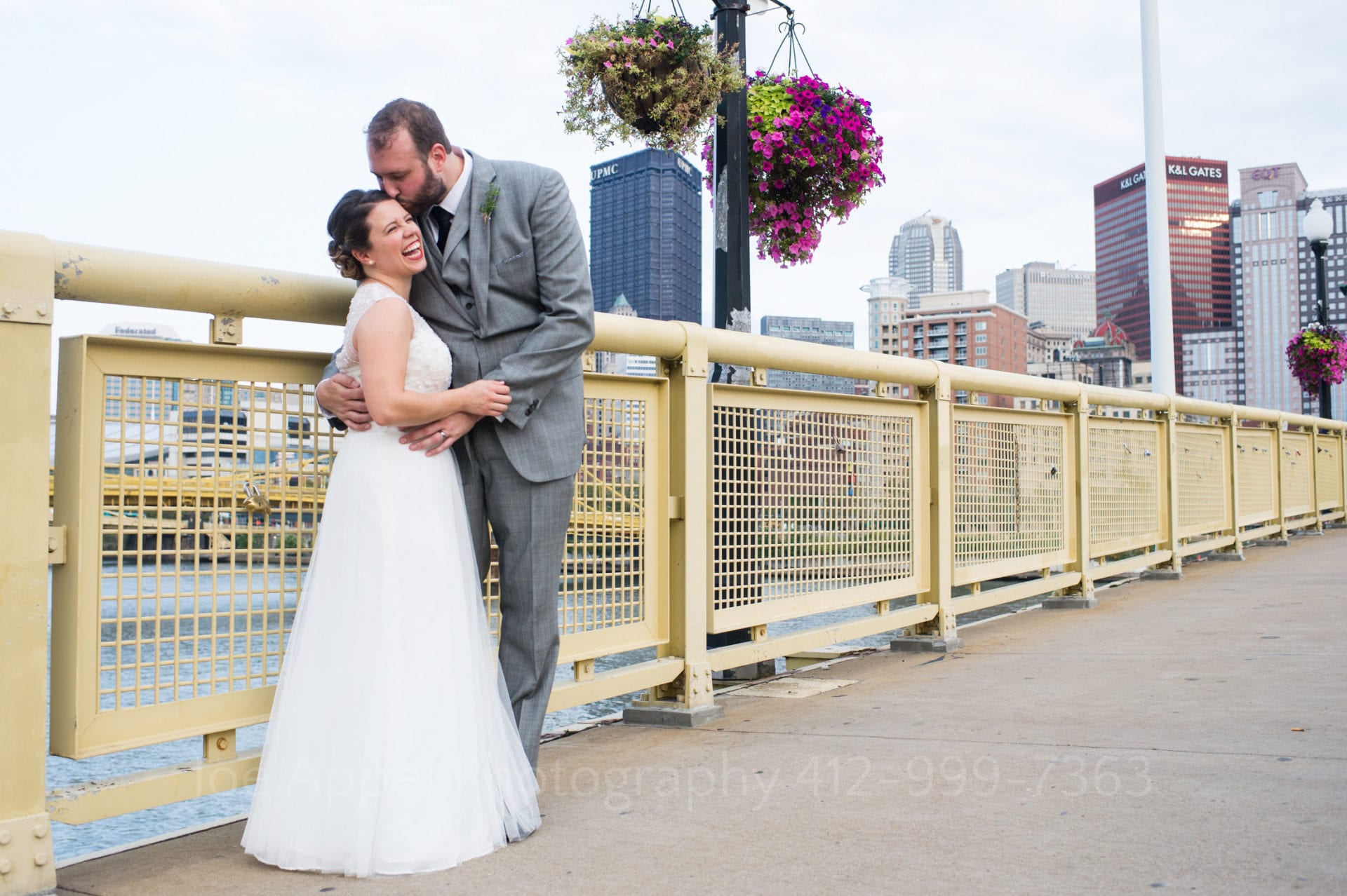 A groom kisses his bride on the forehead as they stand on the walkway of the Roberto Clemente Bridge with the buildings of downtown Pittsburgh behind them.