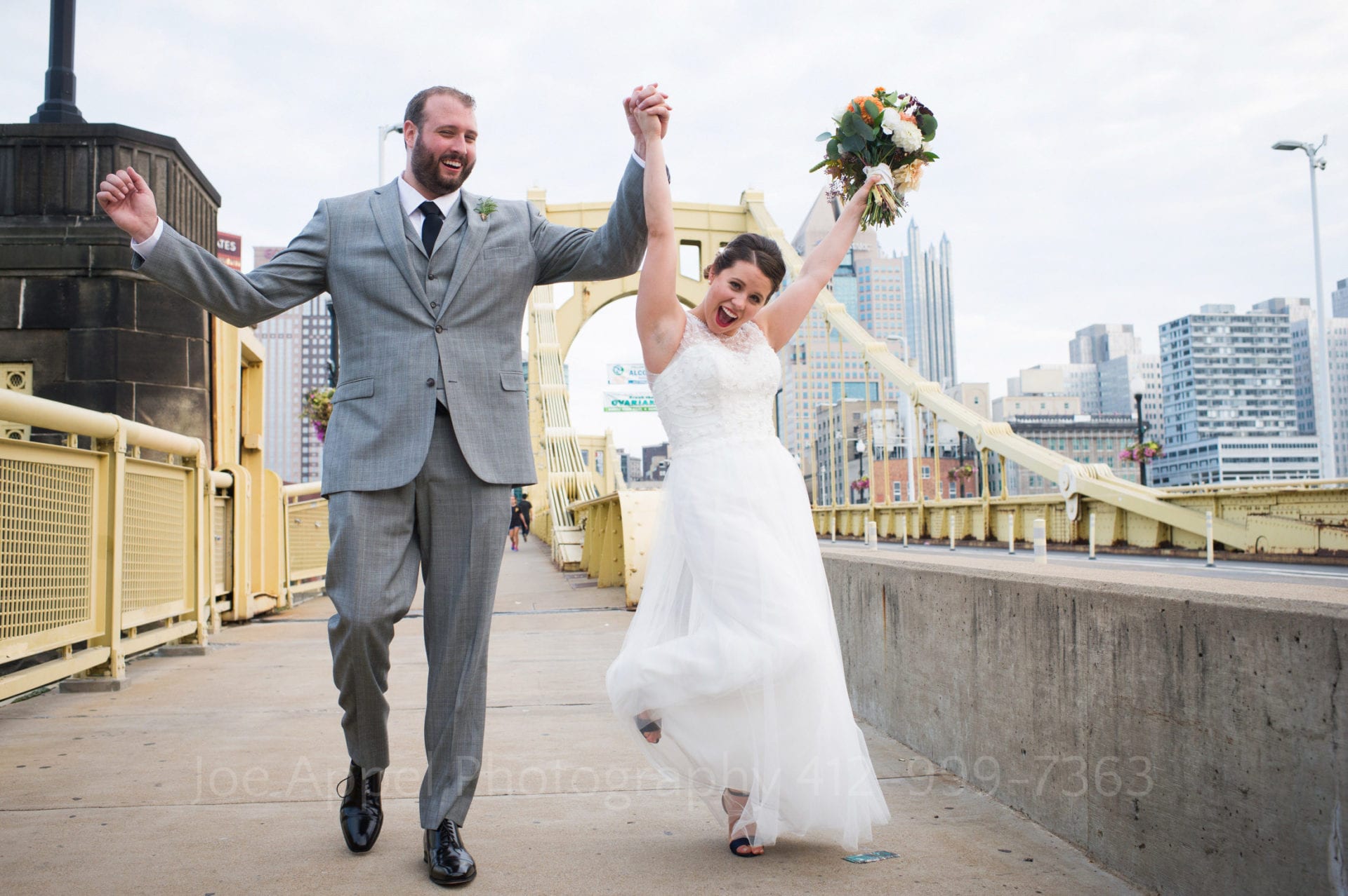 A bride and groom hold their hands high in the air in celebration as they walk across the yellow Roberto Clemente Bridge.