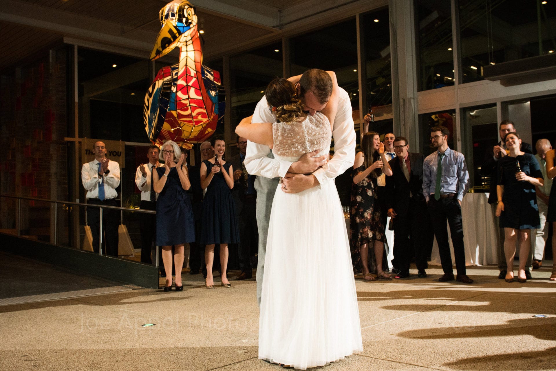 A couple embraces at the end of their first dance while their guests applaud. There is a giant colored glass bird in the background.