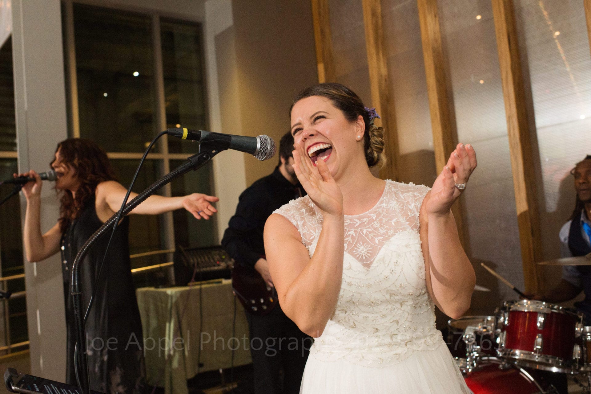 A bride laughs and claps her hands as she sings a song into a microphone.