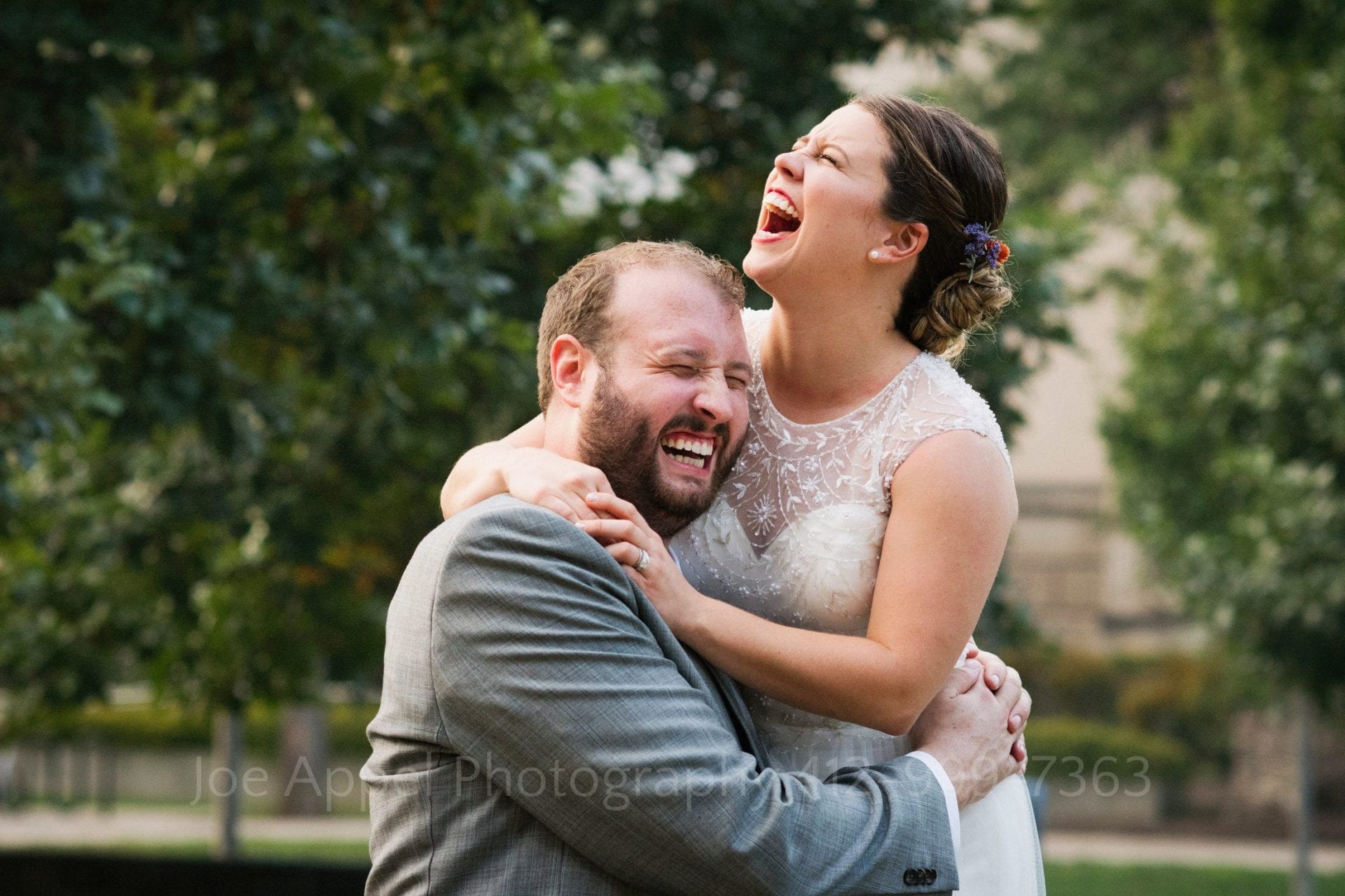 A young bride and groom hold on to one another in a wooded setting and laugh.