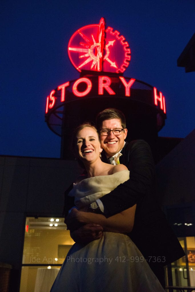 A smiling couple embraces beneath the red neon sign atop of the building during their Heinz History Center wedding.
