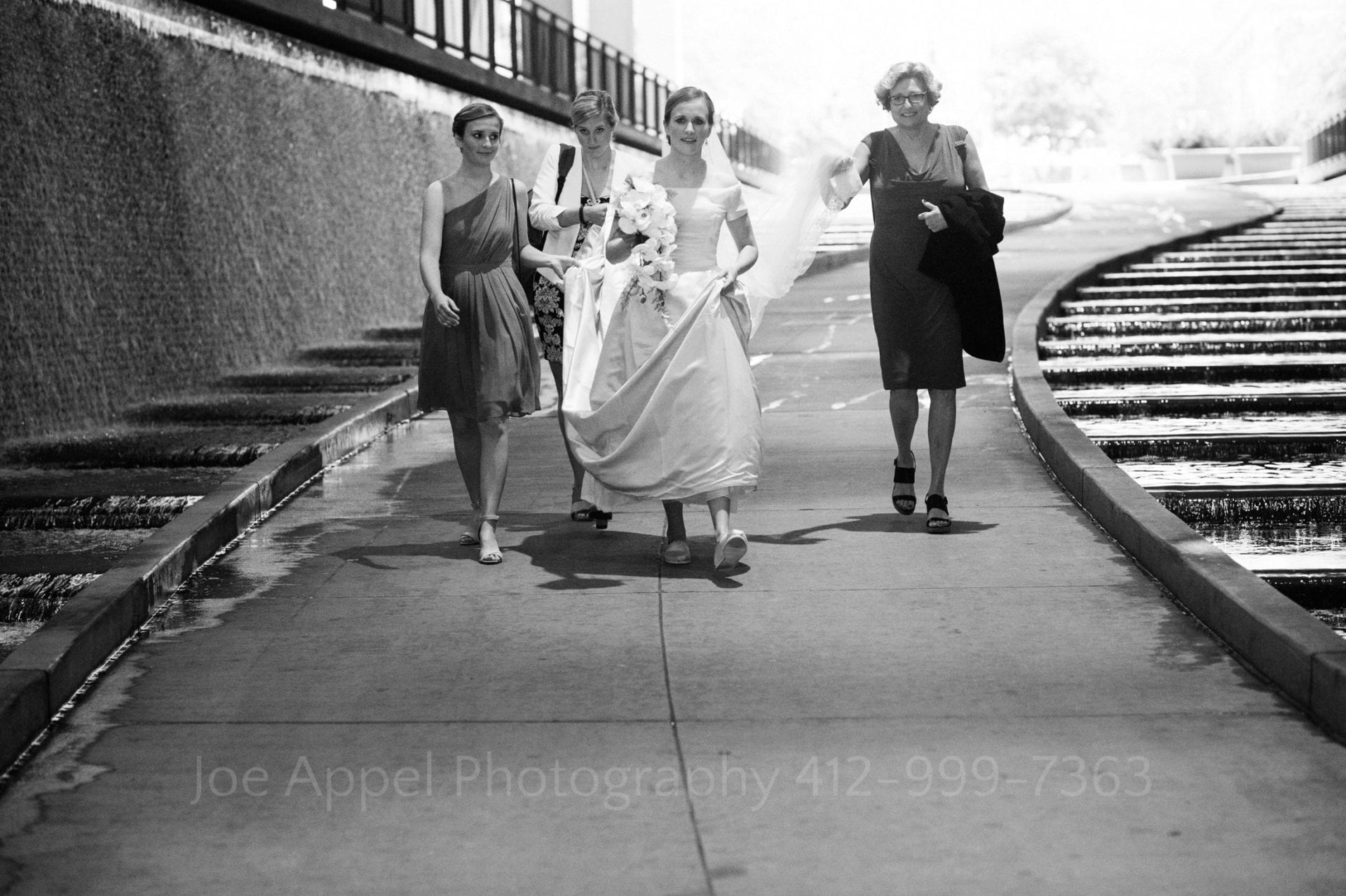 A bride holds her bouquet as she walks down the path beneath the D.L. Lawrence Convention Center with three other women holding her dress.