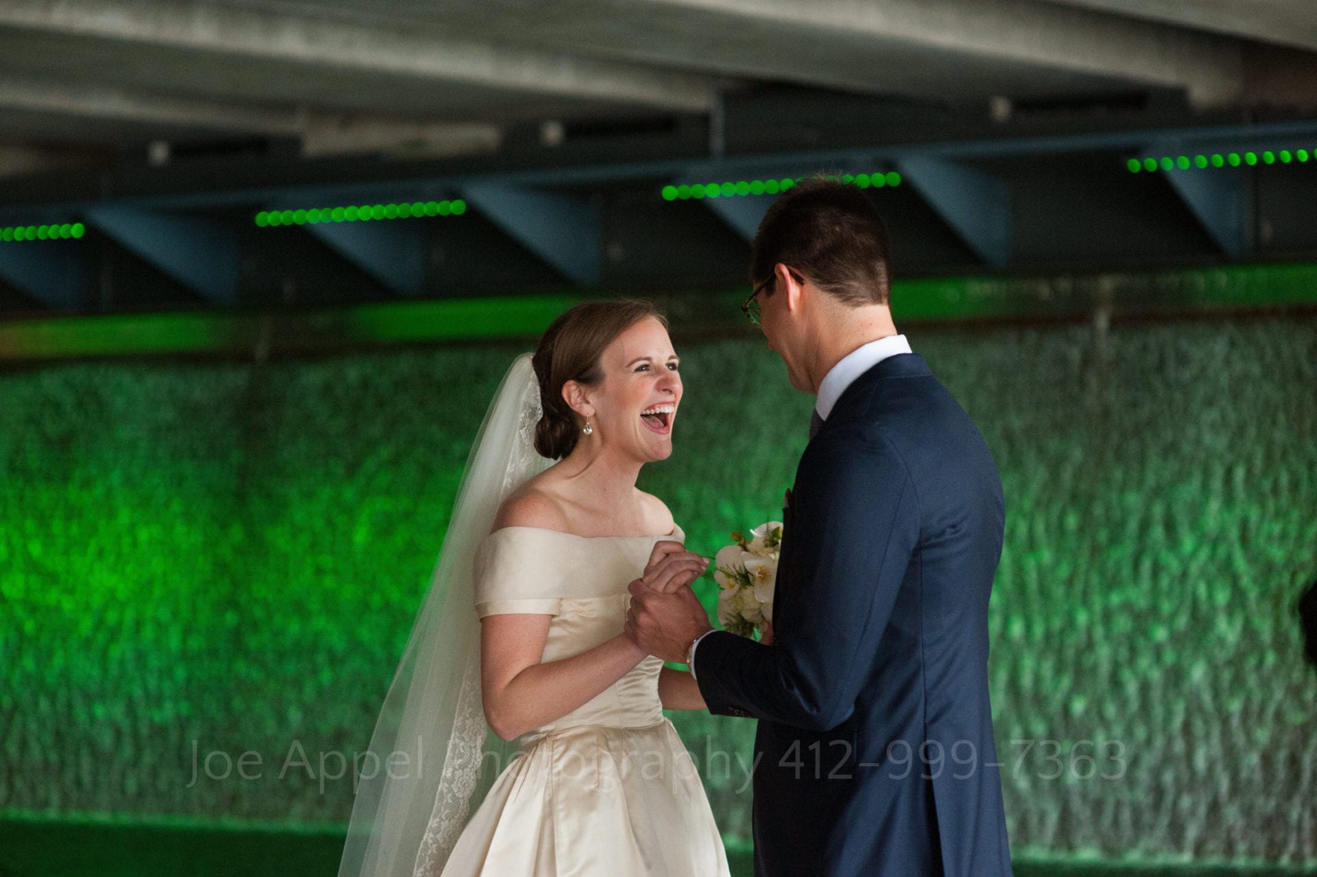 A bride in a white silk dress and veil laughs as she and her groom hold hands while facing each other during their first look. There is a green-lit waterfall behind them.
