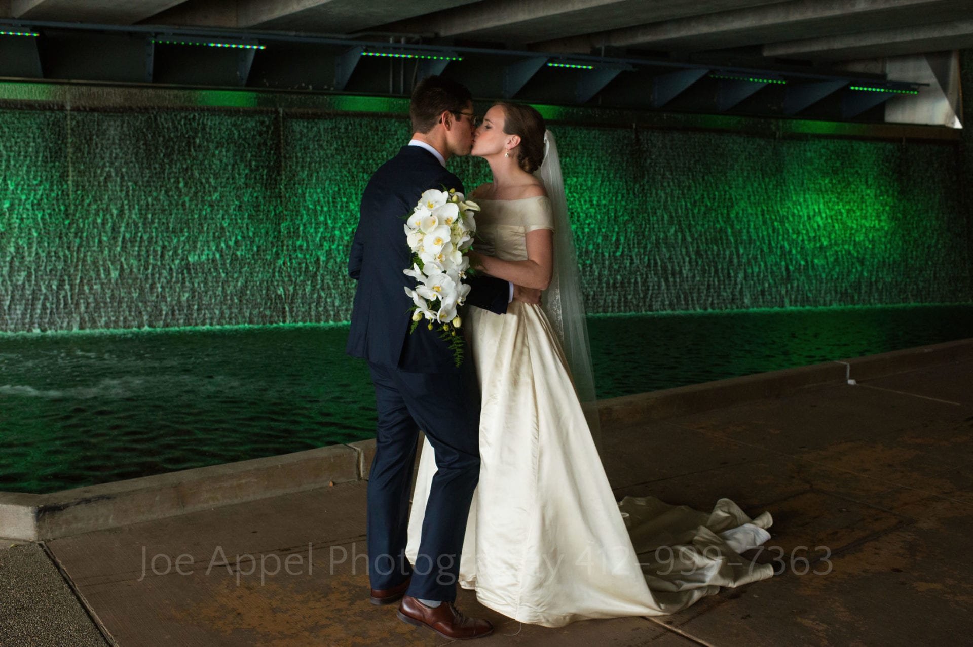 A bride in a white silk dress holds a white bouquet as she kisses her groom in front of a green-lit waterfall.