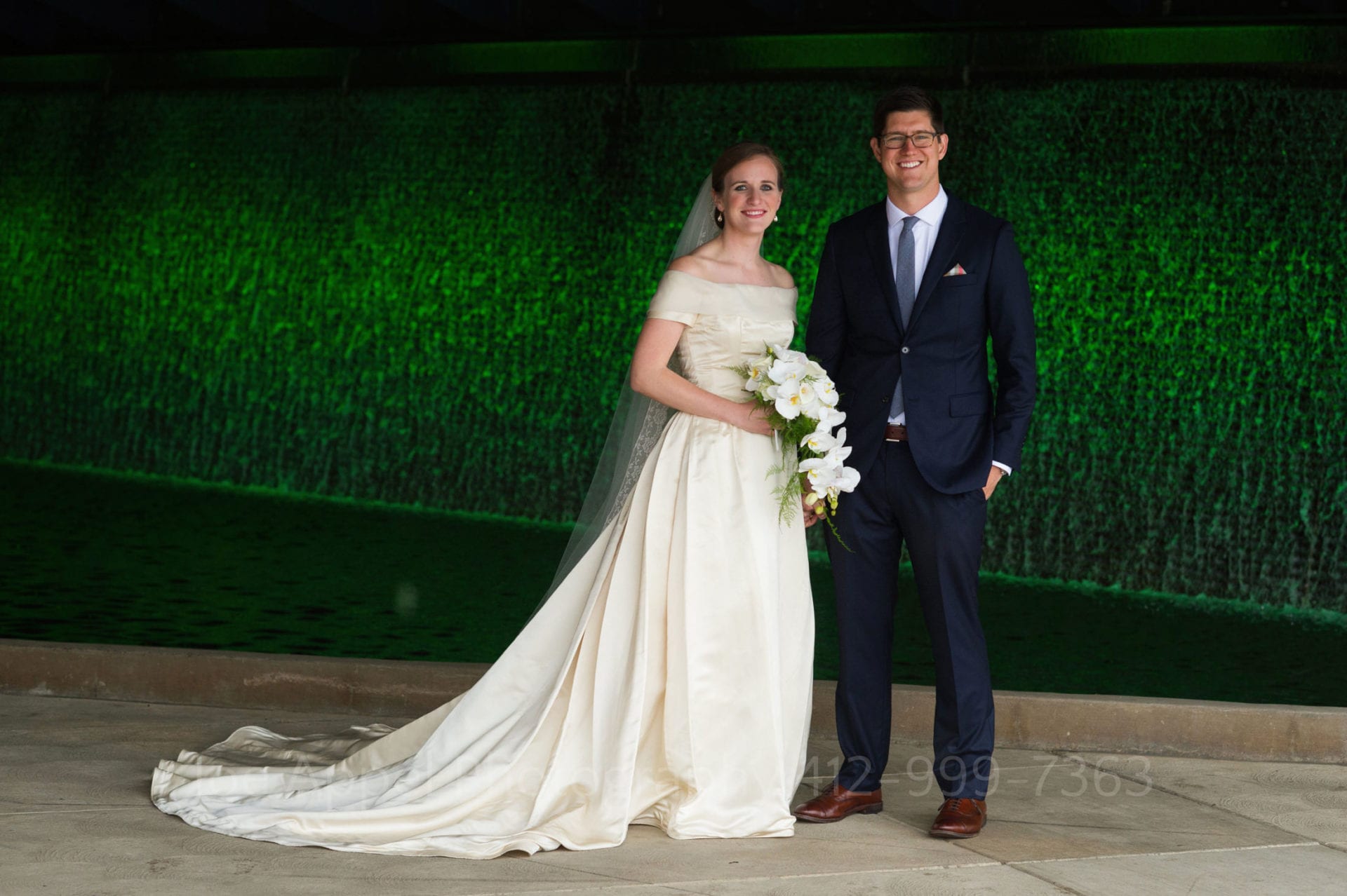 Portrait of a bride and groom standing beneath the D.L. Lawrence Convention Center in front of a green-lit waterfall. The bride wears an off-the-shoulder white silk dress and the groom wears a navy blue suit and brown shoes.
