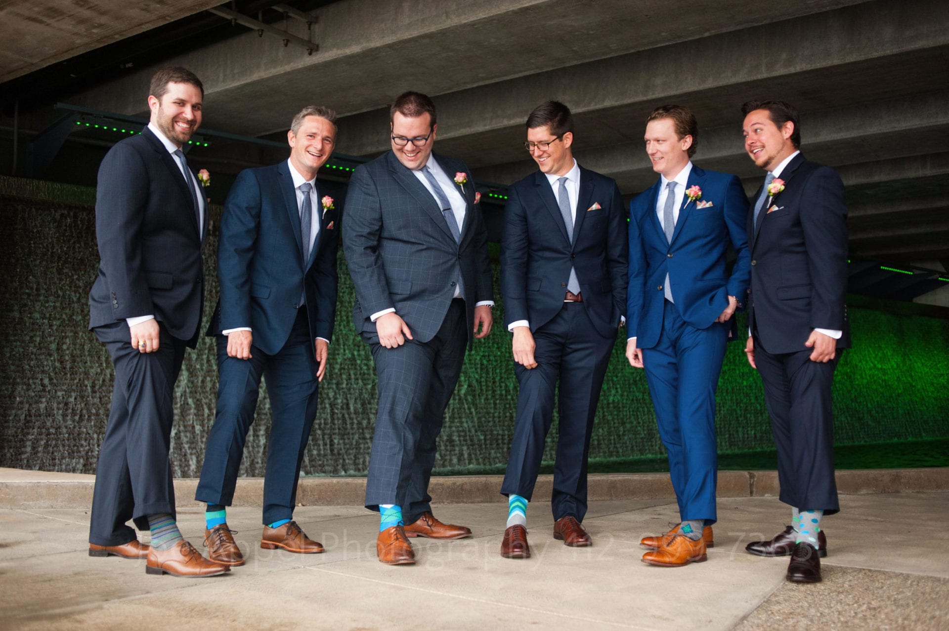 6 men wearing different styles of blue suits hike up their pants legs to show off their bright blue patterned socks.