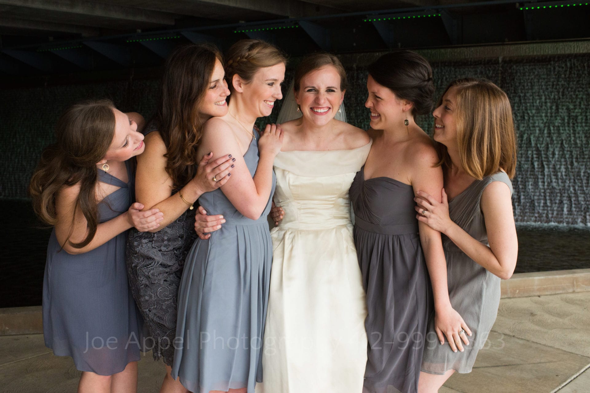 A bride in a white silk dress smiles while hugging her 5 bridesmaids who are dressed in different styles of gray dresses.