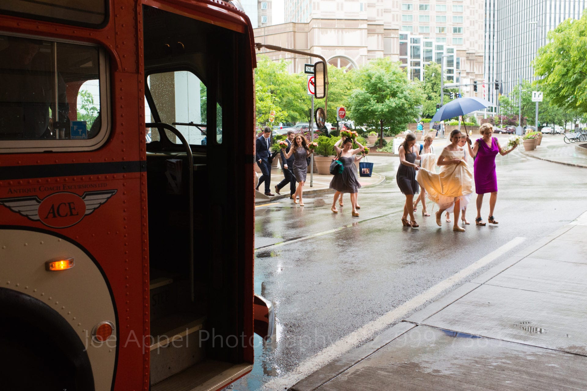 A group of women walk through a rainstorm escorting a bride wearing a white silk dress beneath an umbrella. They're walking to a vintage, orange bus.