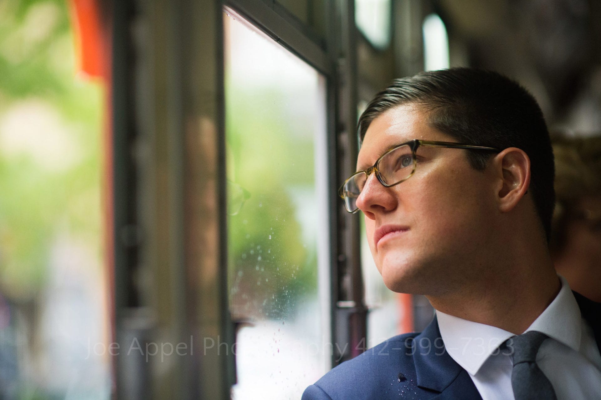 A young man wearing tortoise shell glasses looks out of a rain speckled bus window. There is a raindrop on the shoulder of his blue suit.