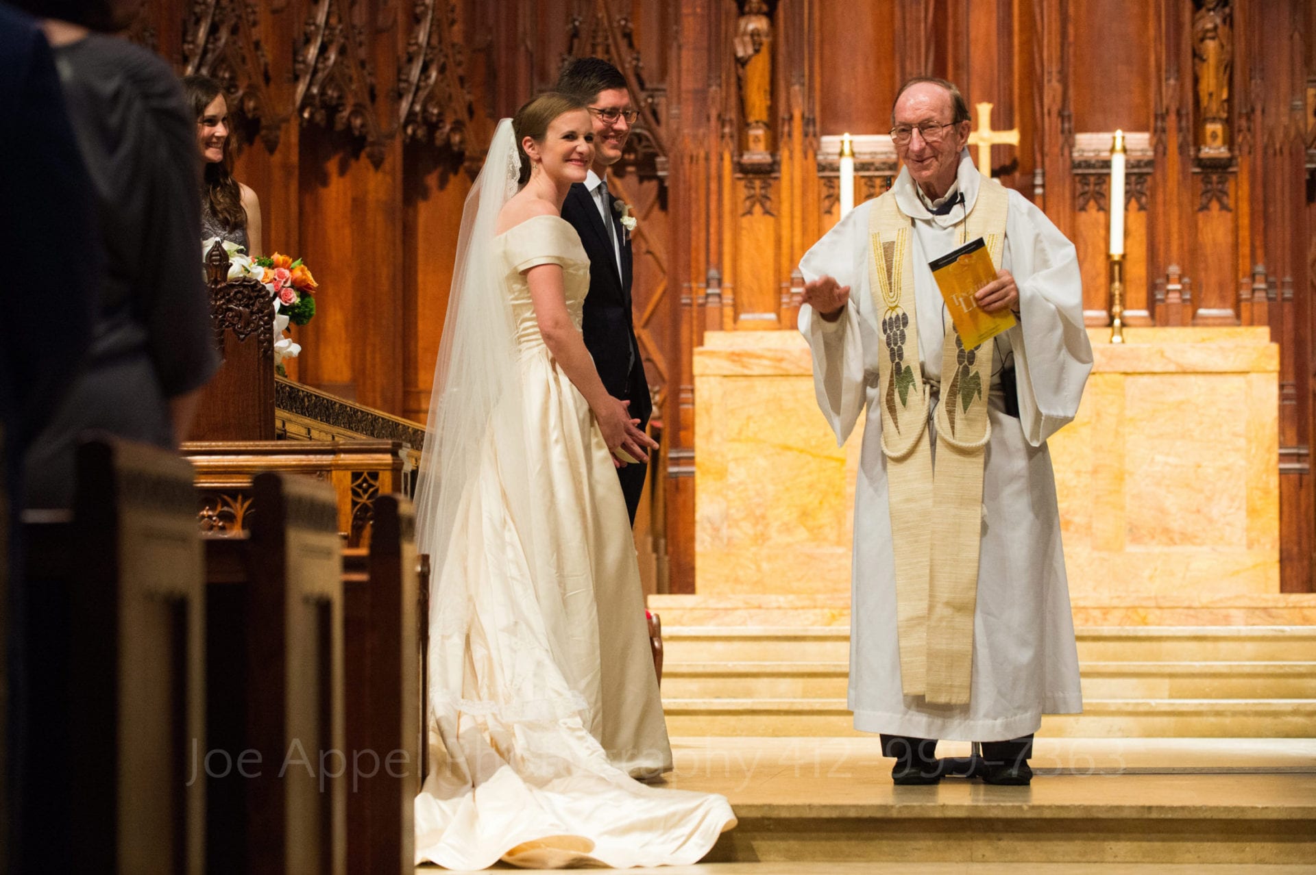 An old priest talks to the congregation as a smiling young bride and groom stand side-by-side at a right angle to him on the altar during their Heinz Chapel wedding.