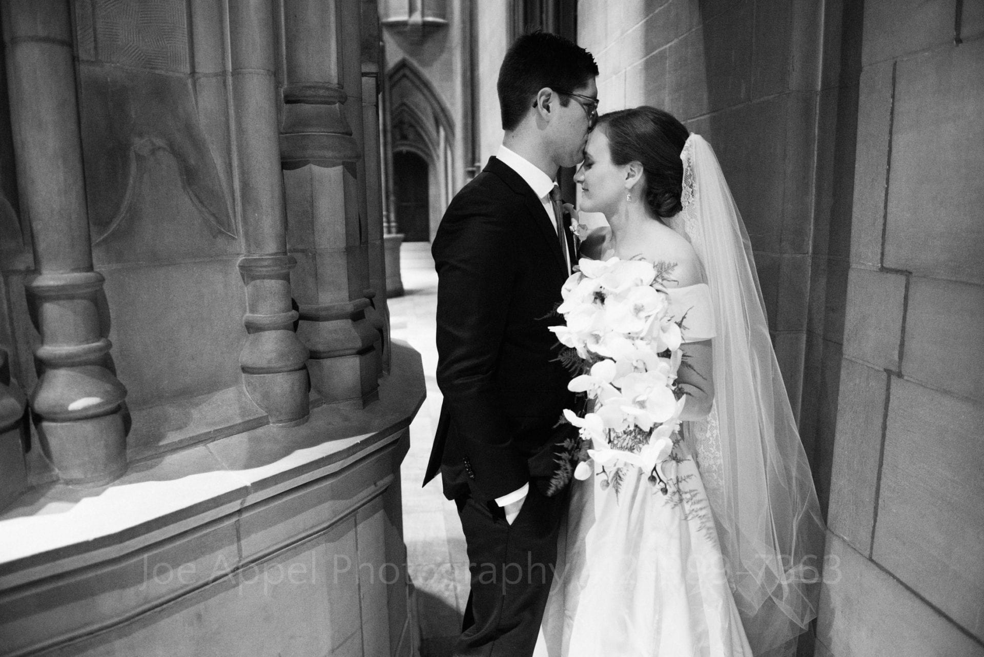 A groom kisses his bride on the forehead while they stand behind a stone column at the side of the church during their Heinz Chapel Wedding.