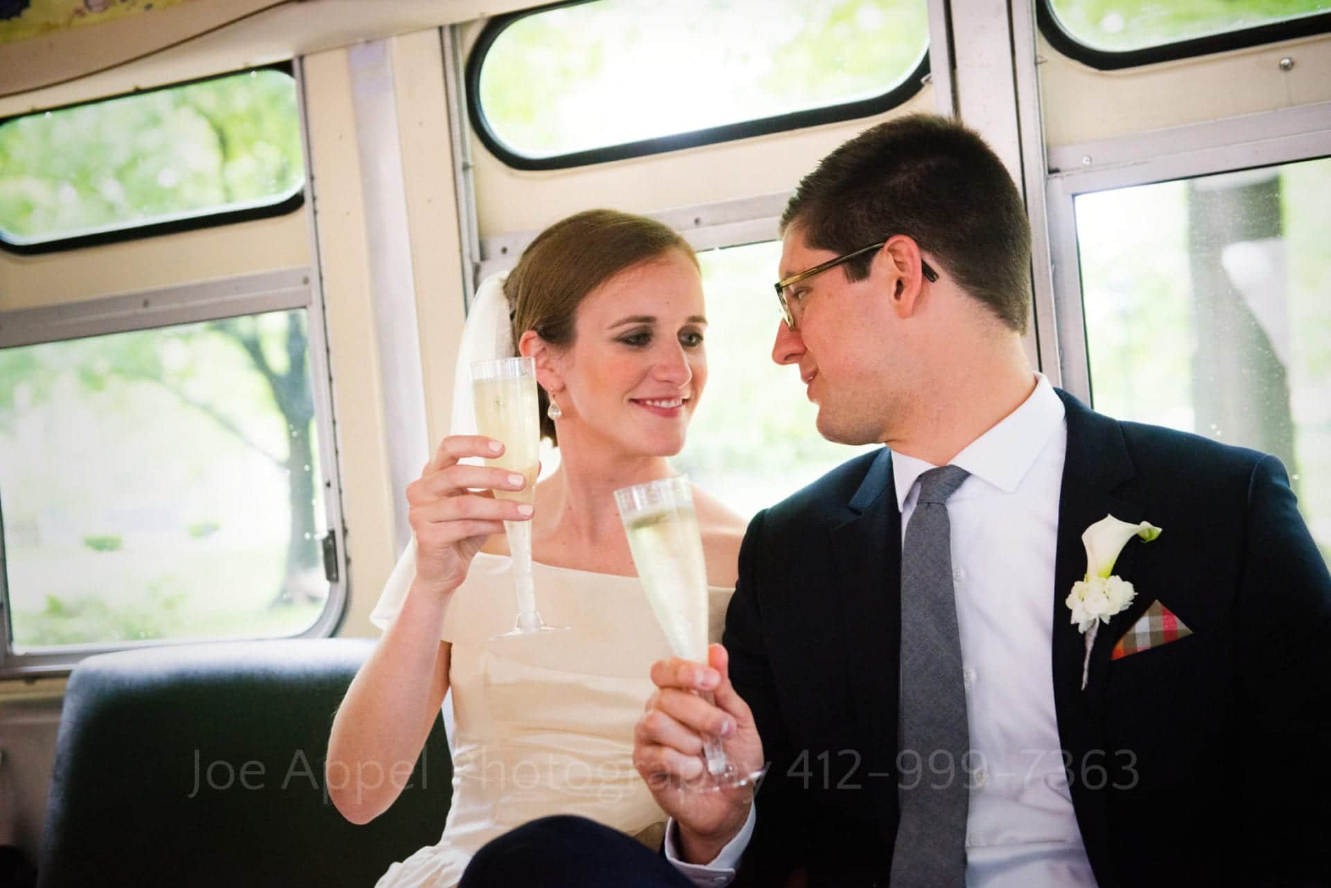 A bride and groom toast with plastic flutes filled with champagne as they sit side-by-side on a vintage bus.