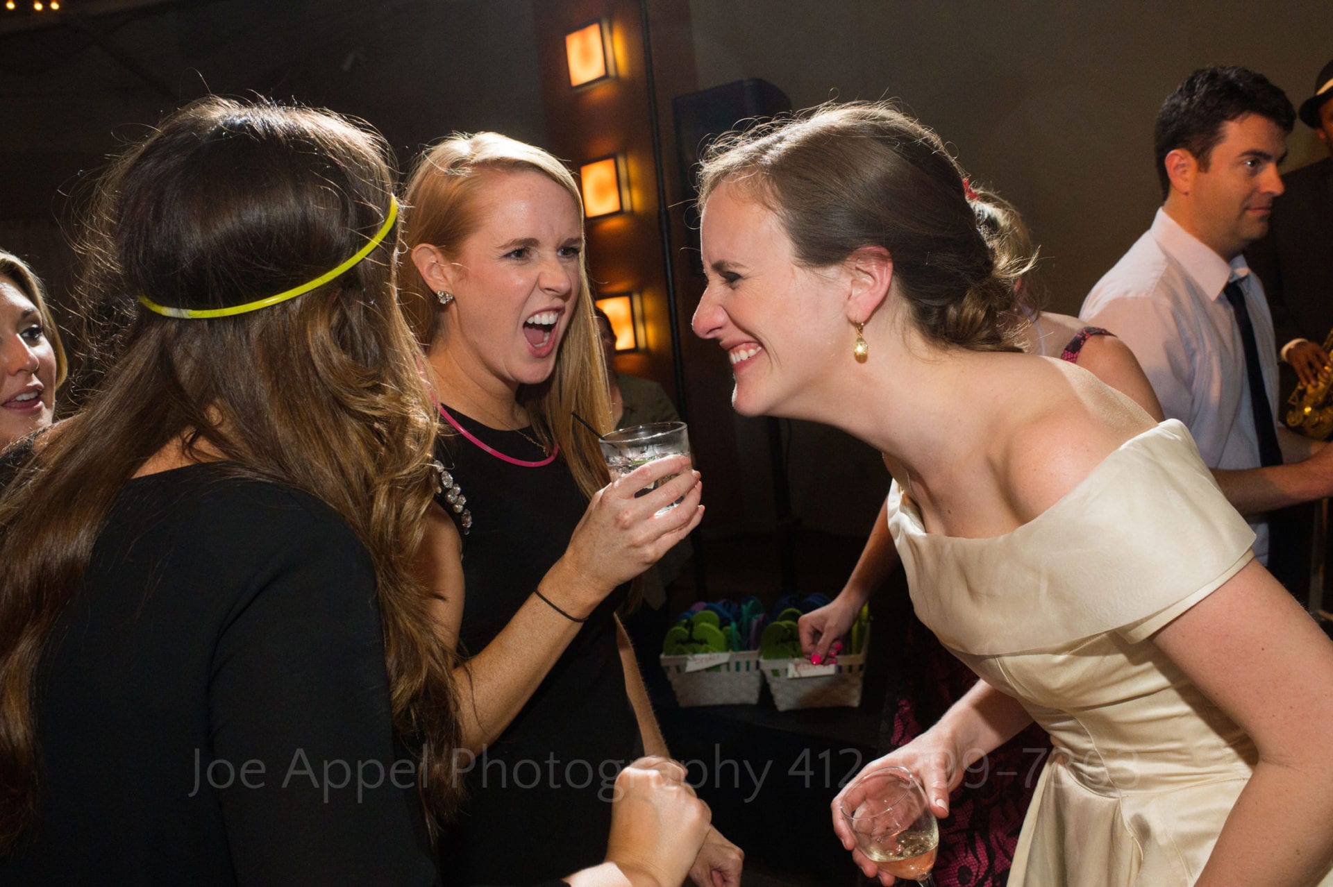 A bride smiles as she dances with two female friends one of who is yelling and holding a glass of liquor during a Heinz History Center wedding
