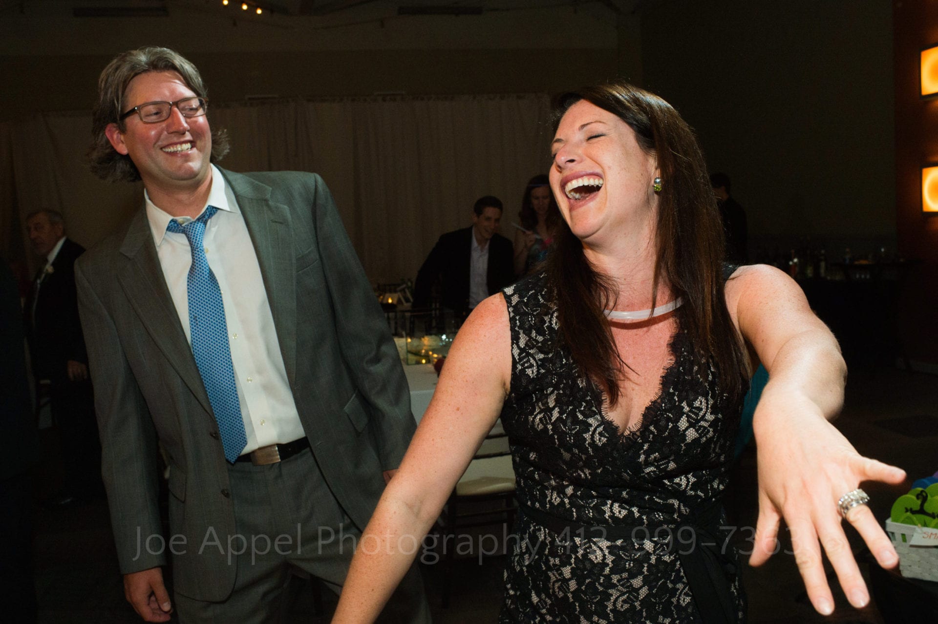 A woman in a black lace dress holds out her hand and laughs while dancing with a man in a gray suit and blue tie.