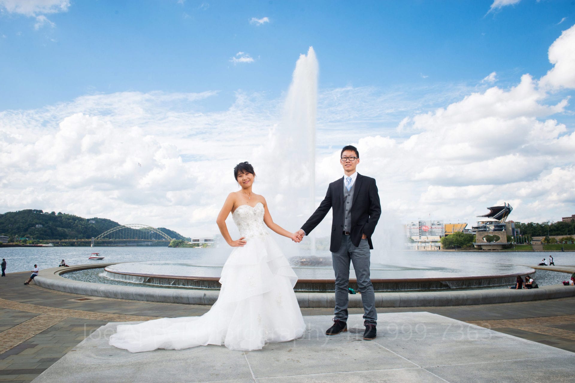 a bride and groom hold hands as they smile and pose in front of the point state park fountain