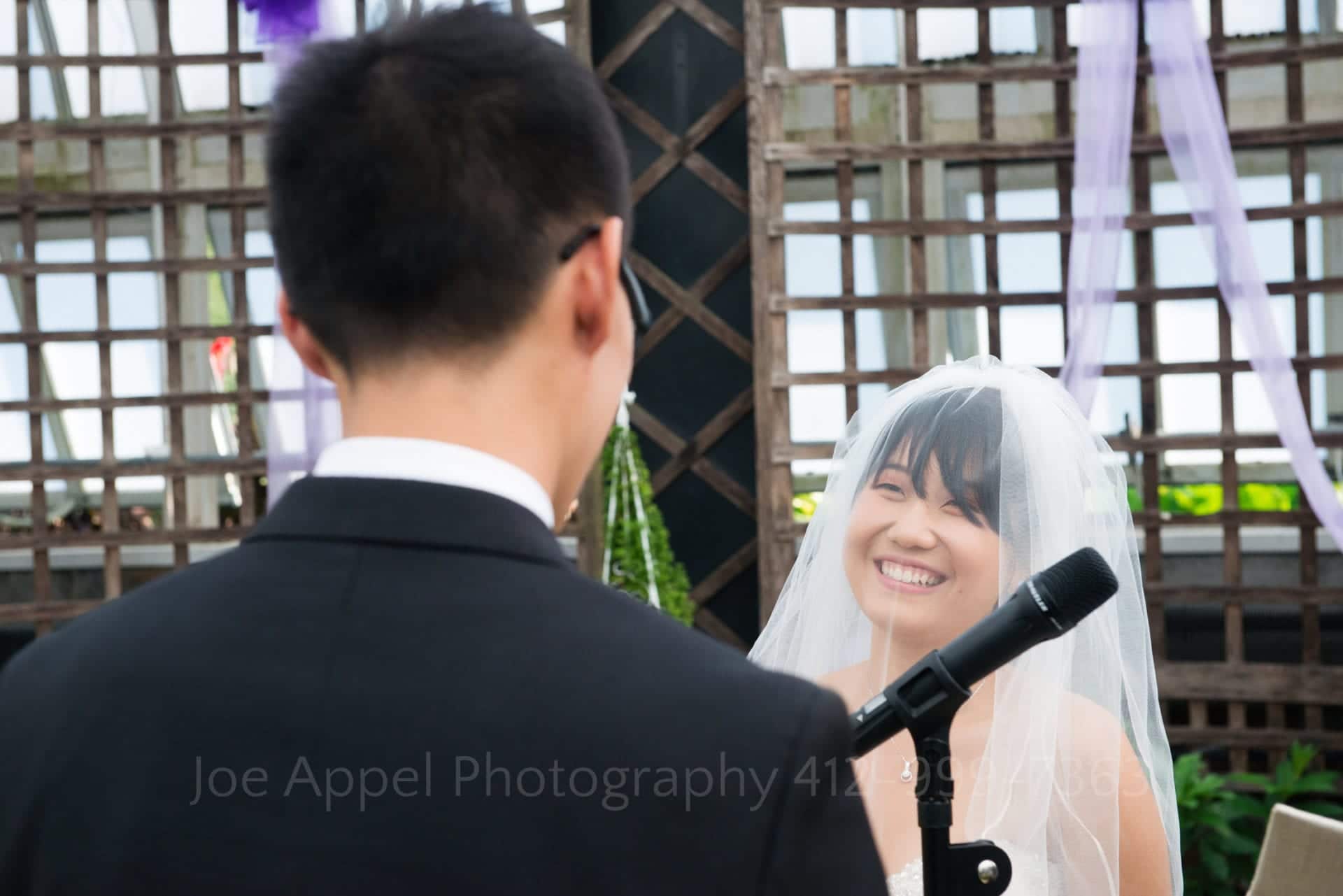 a bride smiles at her groom as they face each other during their ceremony in the Broderie Room at Phipps Conservatory wedding.