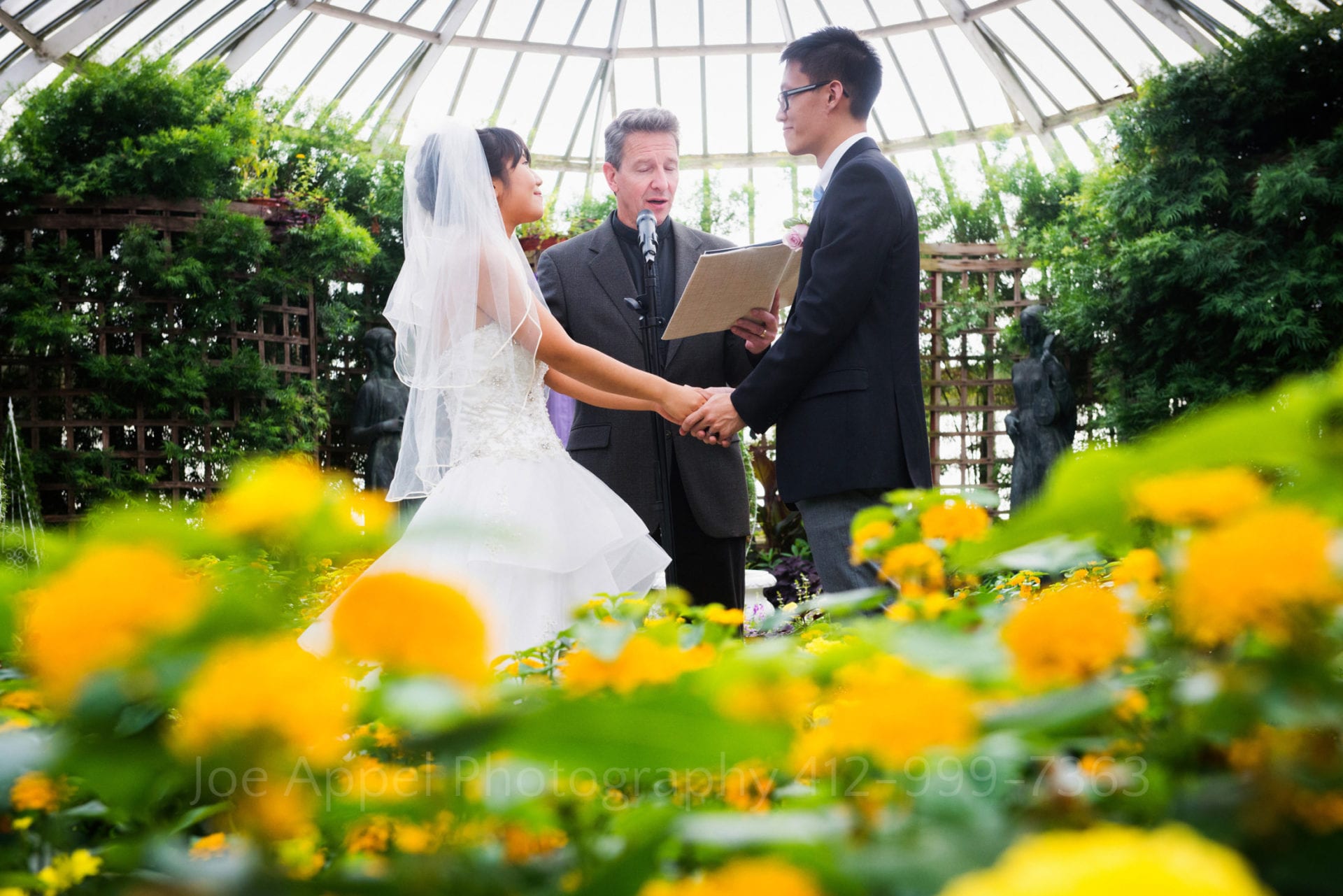 a bride and groom say their vows surrounded by yellow flowers in the broderie room during their Phipps Conservatory wedding.