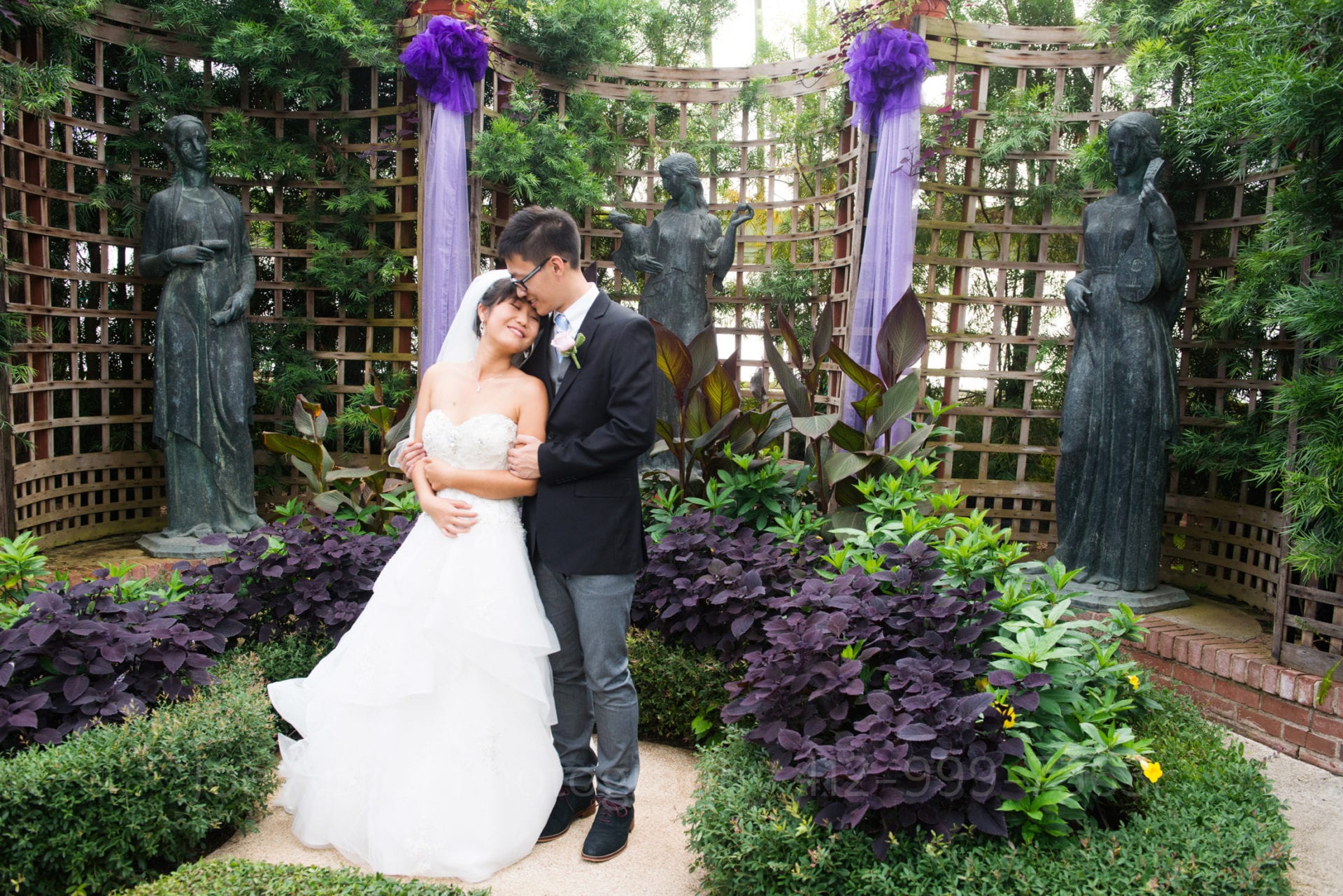 a bride in a white dress smiles with closed eyes while leaning into her groom. There are statues of women in the background with purple bunting hanging off of a trellis.