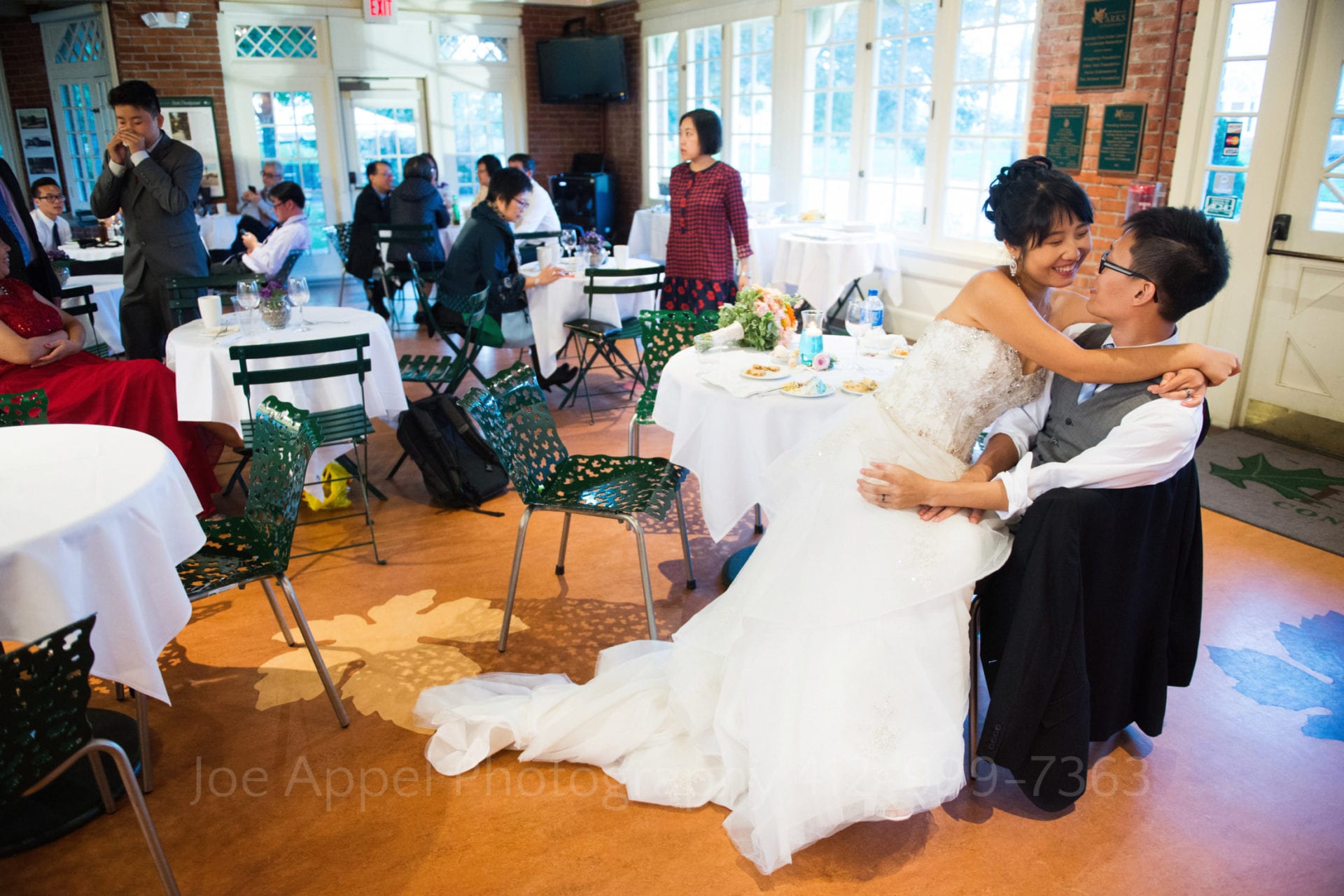 a bride sits on her grooms lap and smiles at him during their wedding reception at the schenley park cafe and visitors center.