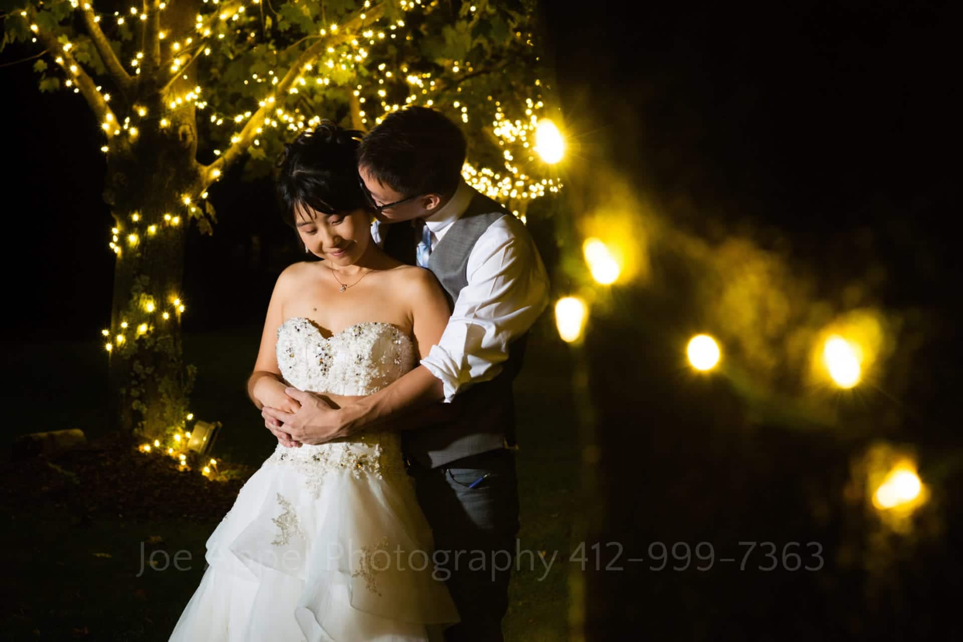 a bride and groom embrace in front of golden fairy lights during their Grand Estate at Hidden Acres wedding.