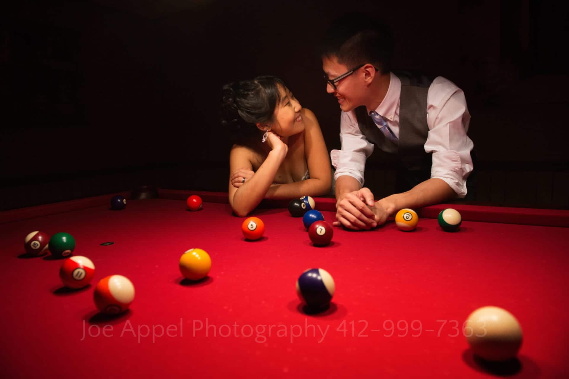 a bride and groom lean on a pool table with a red felt top and smile at each other during their grand estate at hidden acres wedding.