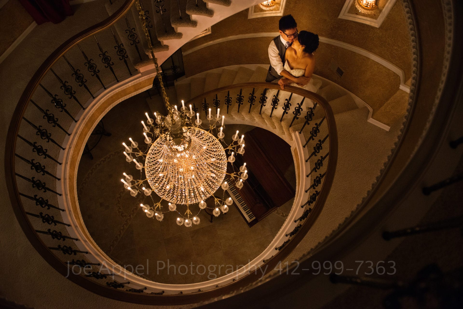 a bride and groom embrace on a spiral staircase next to a chandelier during their grand estate at hidden acres wedding.