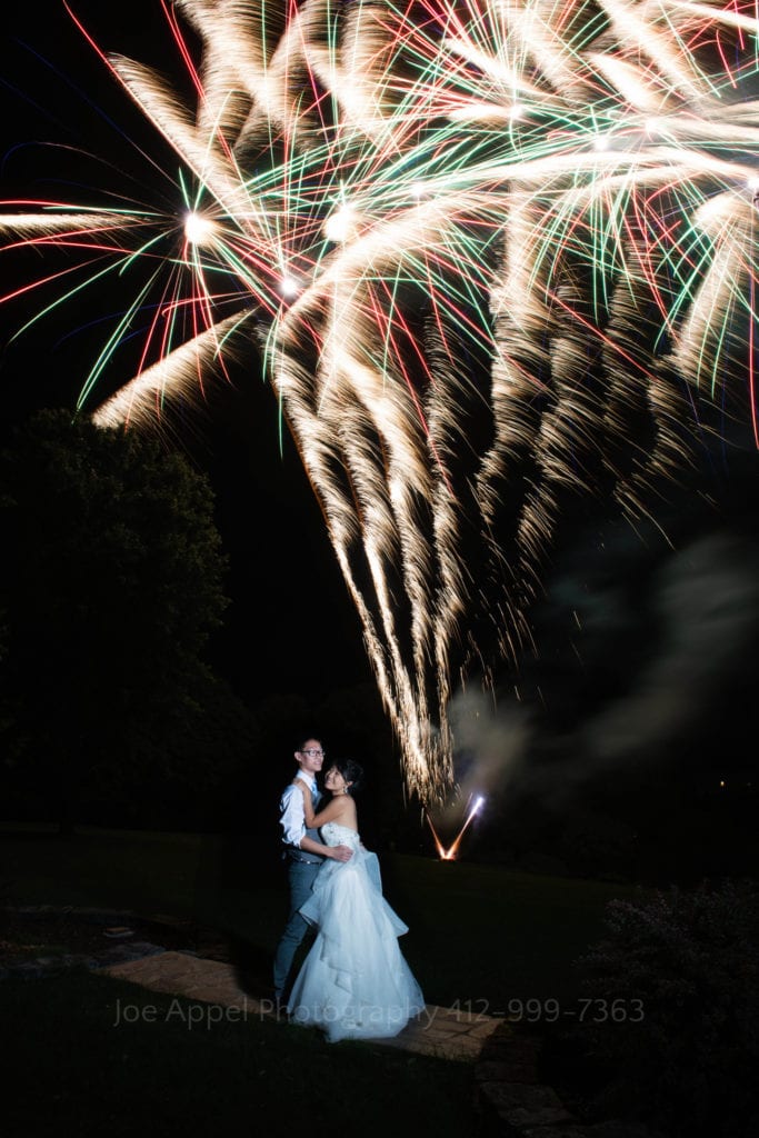a bride and groom embrace in front of red and green fireworks at the end of their grand estate at hidden acres wedding.