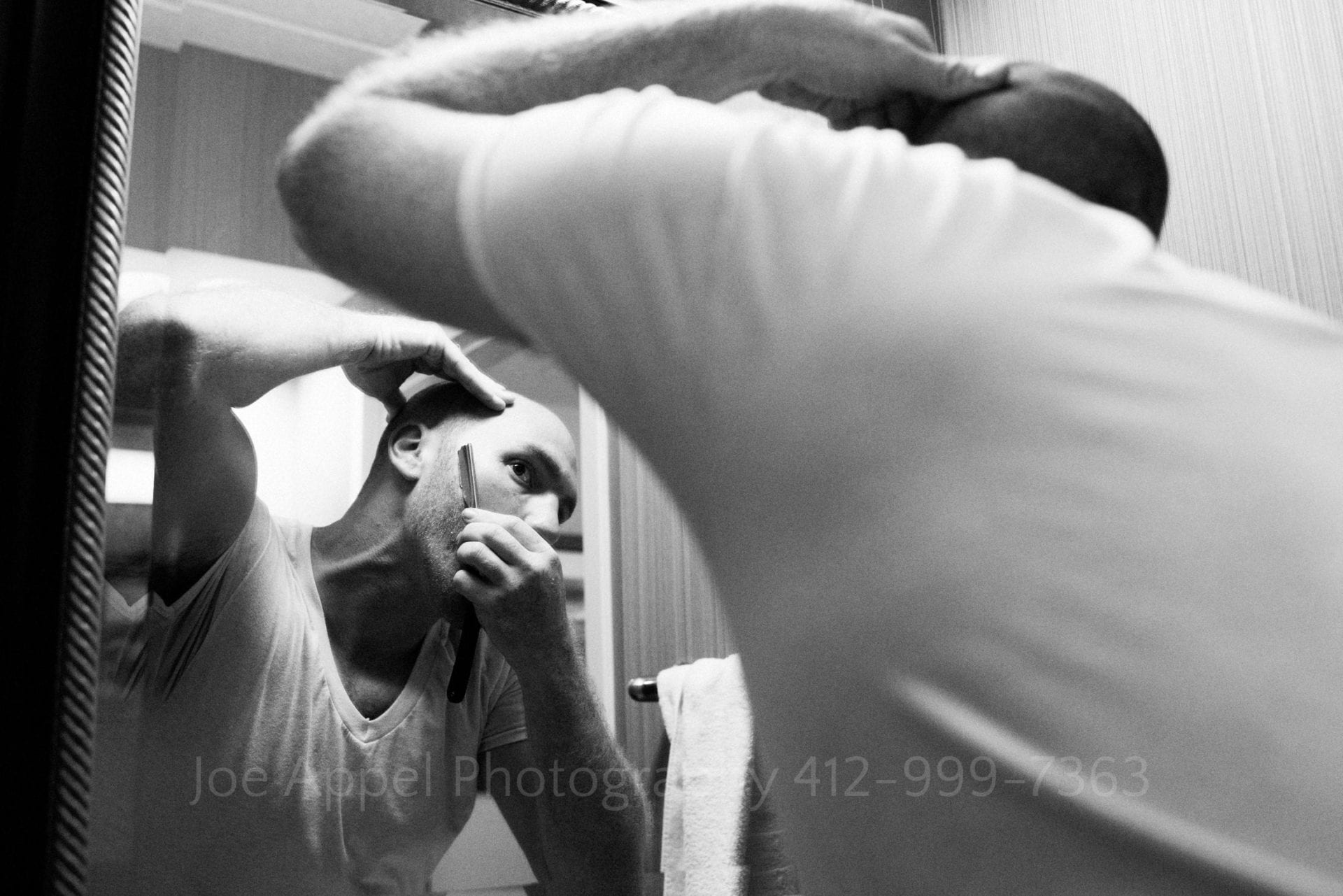 A man wearing a v-neck t-shirt looks in a mirror as he trims his beard with a straight razor.