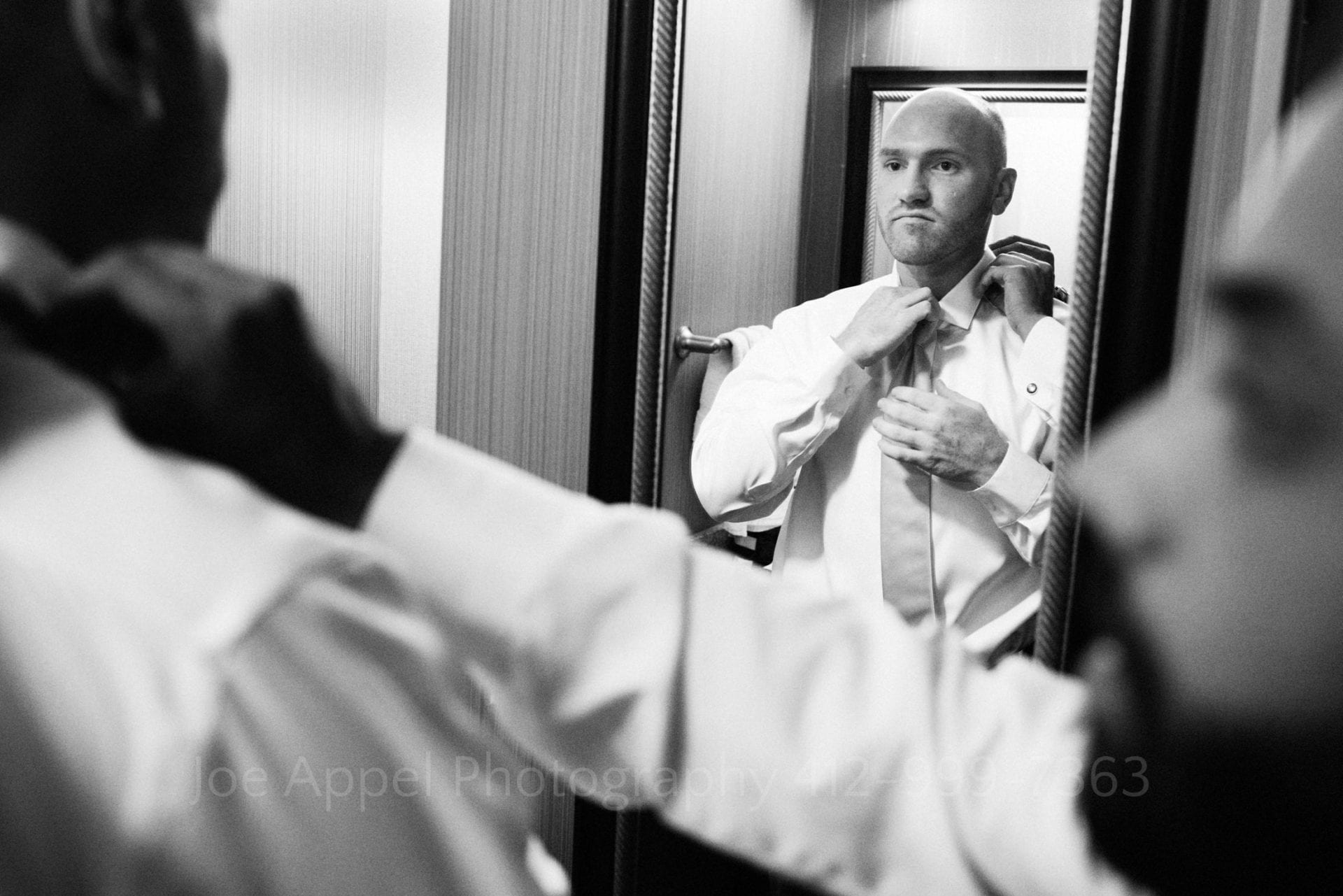 A groom with a shaved head adjusts his tie in a mirror as a friend fixes his collar for him.