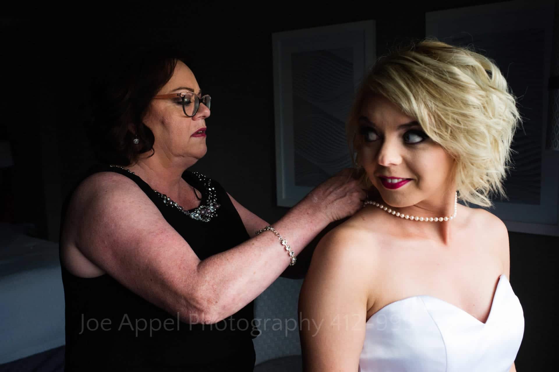 A blonde bride looks back at her mother as her mother clasps a pearl necklace around her neck.