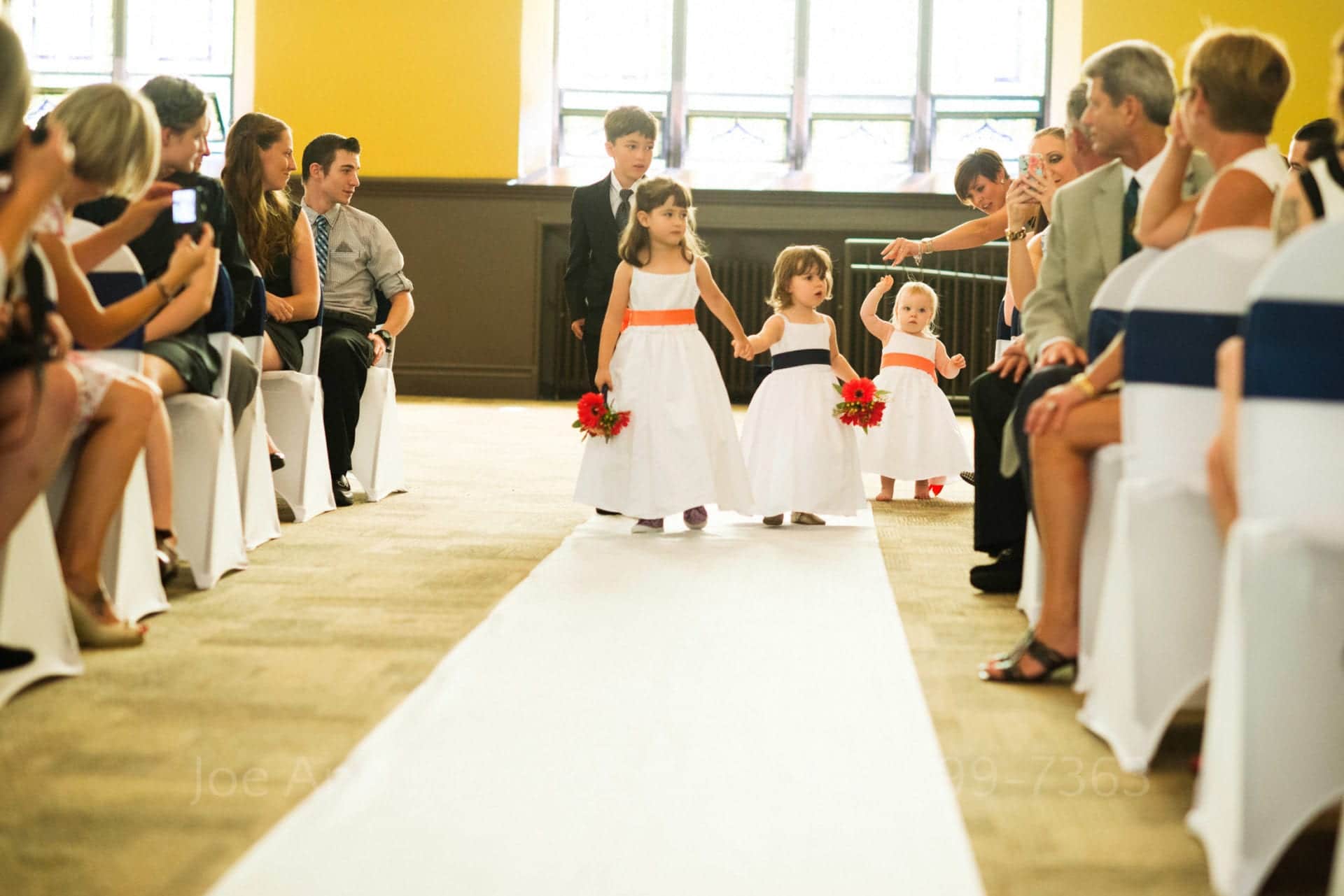Three little girls in white dresses and a boy in a suit are encouraged to walk down the aisle at a wedding.