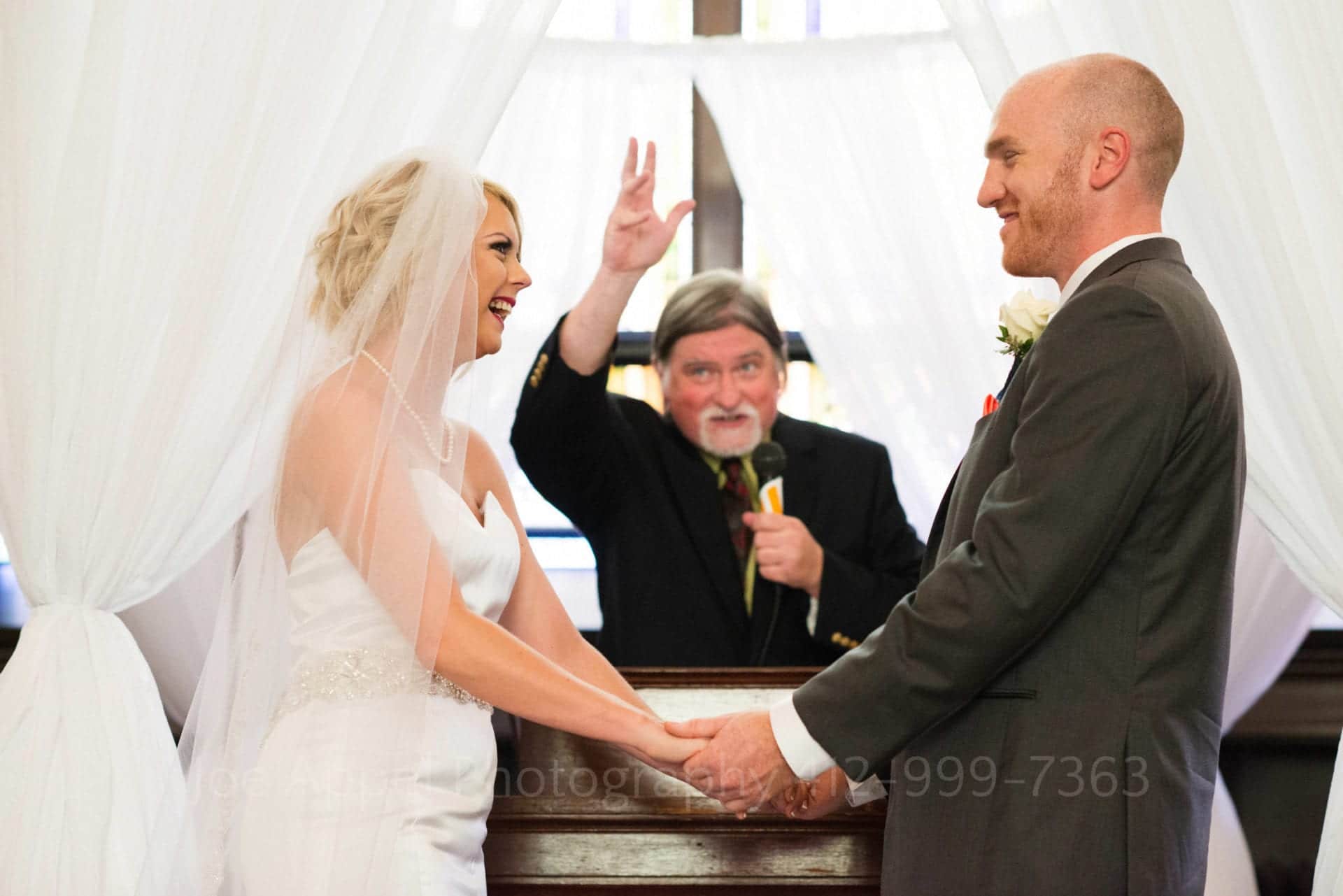 A bearded officiant raises his hand in a blessing between a bride and groom who smile and hold hands during a wedding at the Union Project in Highland Park.