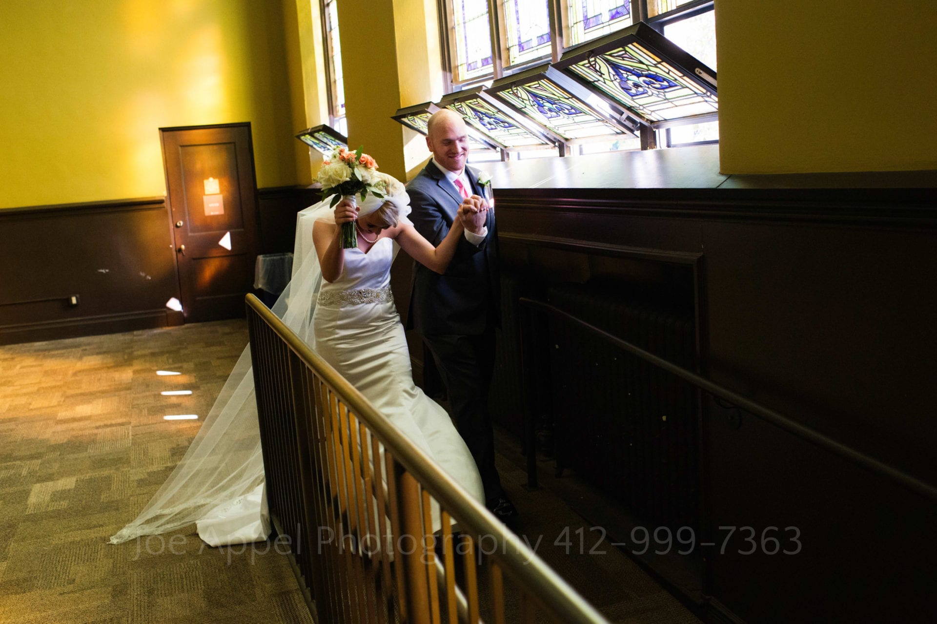 A bride and groom hold their hands up in celebration as they walk down a ramp near some stained glass windows in the big yellow room at the Union Project in Highland Park.