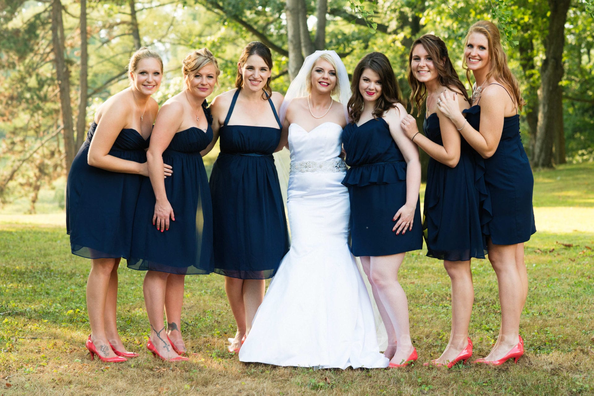 A bride stands with her bridesmaids in a wooded lot as they smile at the camera. The bridesmaids wear navy blue dresses and red shoes.