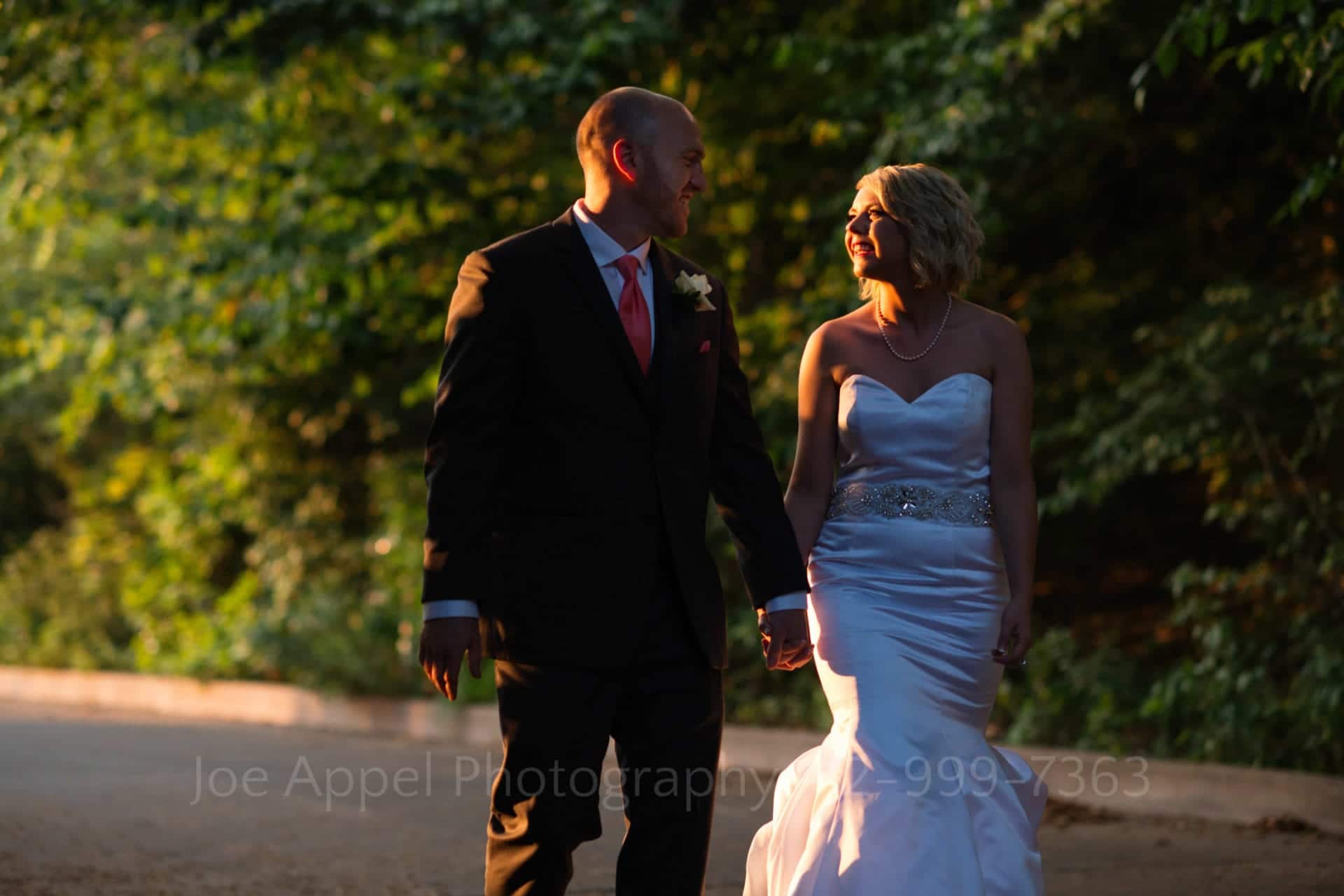 A bride and groom hold hands and walk while illuminated by the setting sun.
