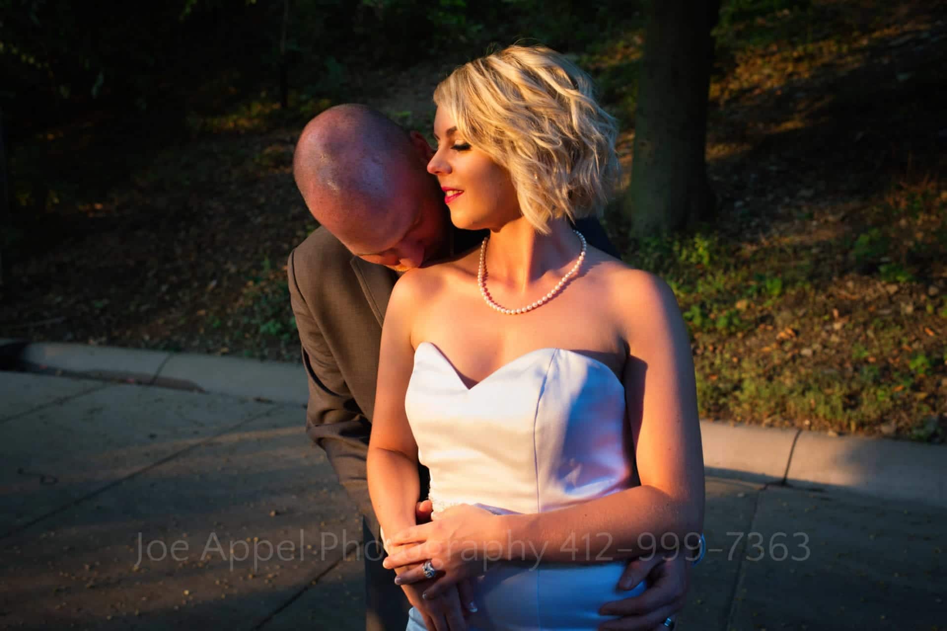 A groom stands behind his bride and kisses her shoulder in the setting sun. She looks to the side and smiles.