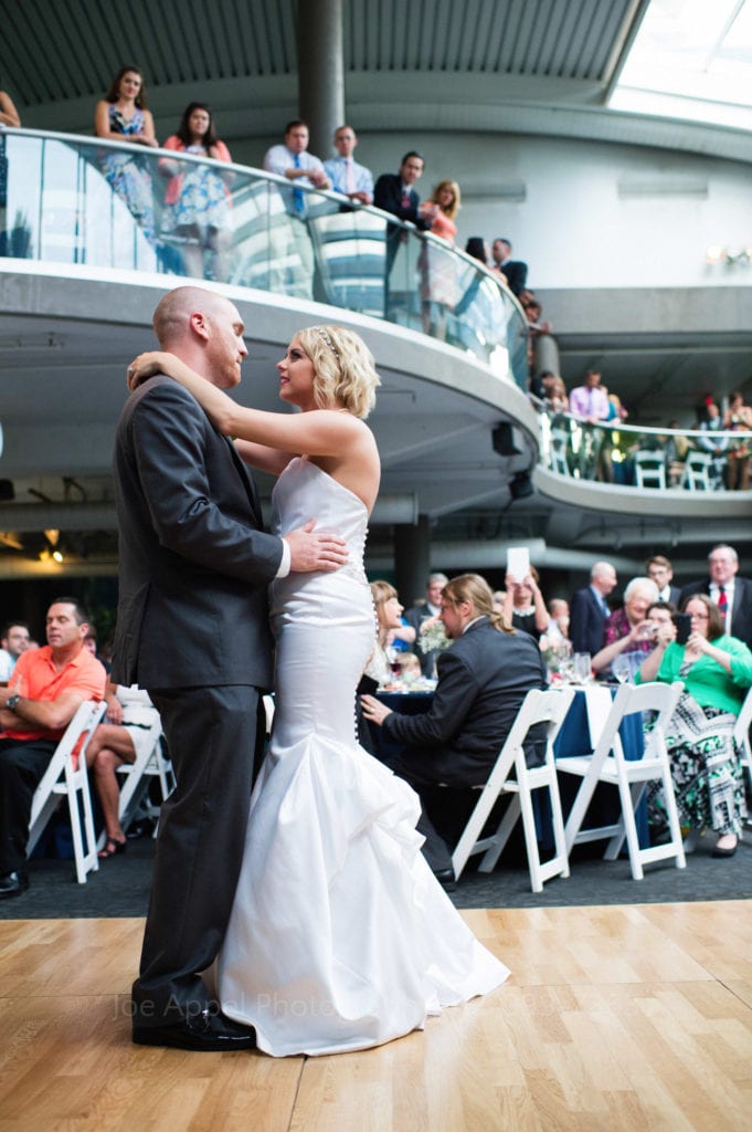 A bride and groom dance in the foreground at their wedding at PPG Aquarium. Their guests watch from the floor and from the curved balcony above.