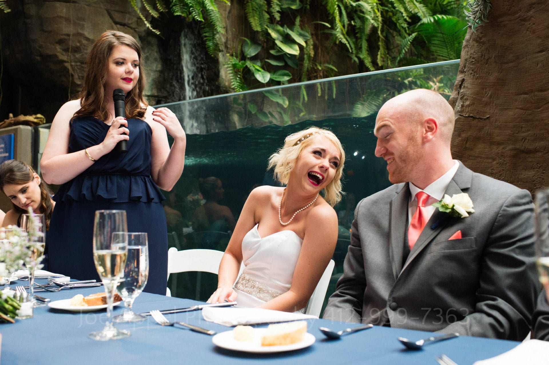 A bride looks at her groom and laughs as her maid of honor gives a speech at their PPG Aquarium Wedding. The groom wears a gray suit with a pink tie. The maid of honor wears a navy blue dress. The bride wears a strapless white gown. There are plates of bread on the blue tablecloth in front of them.