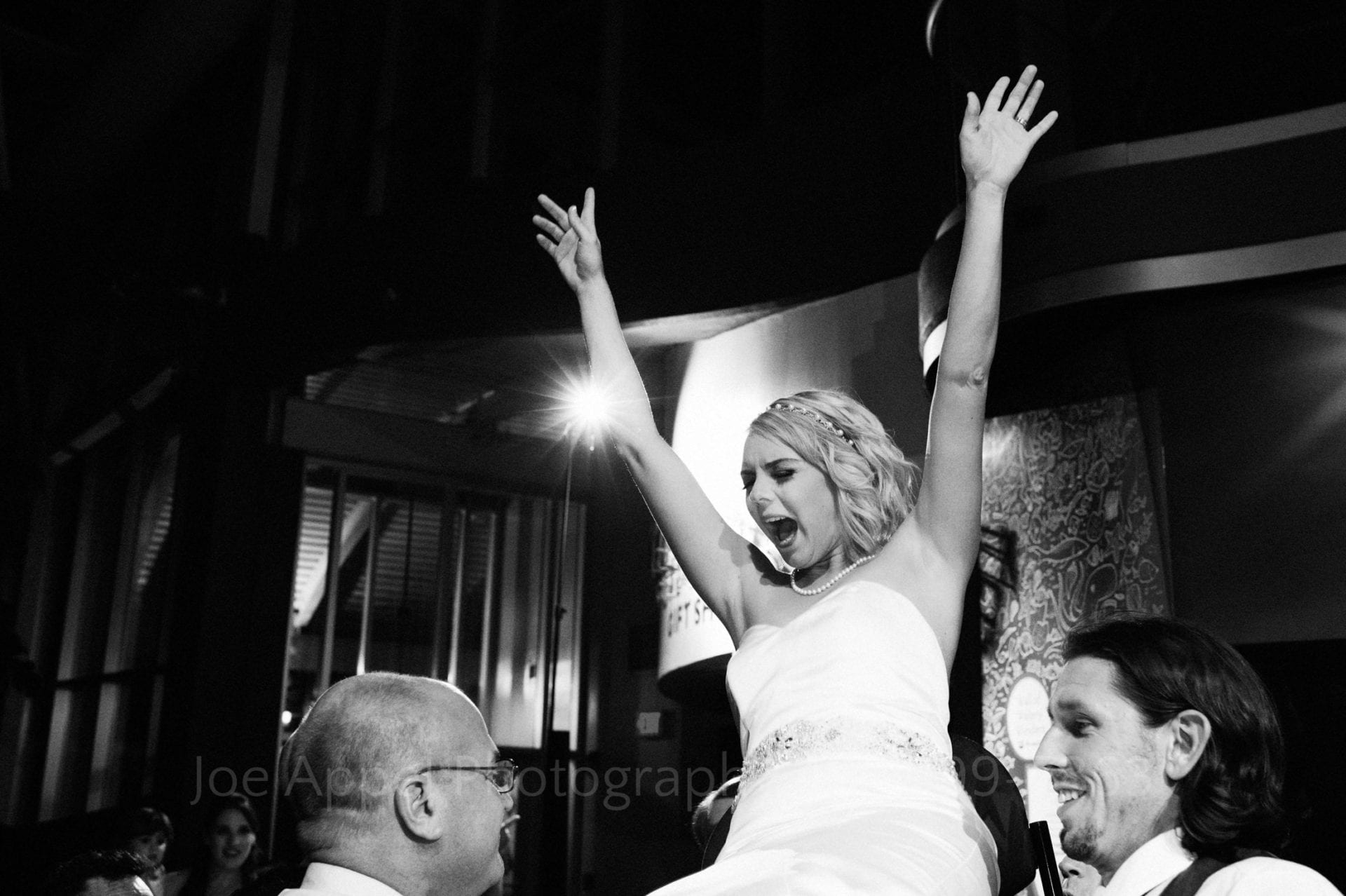 A bride holds her hands in the air and yells as she is held high above the dance floor by two men.