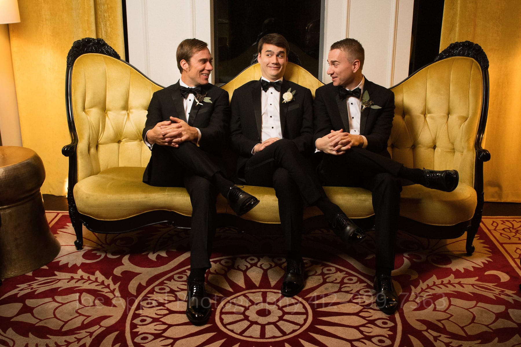 a groom sits crosslegged on a yellow couch with his brothers as they make funny faces at each other.