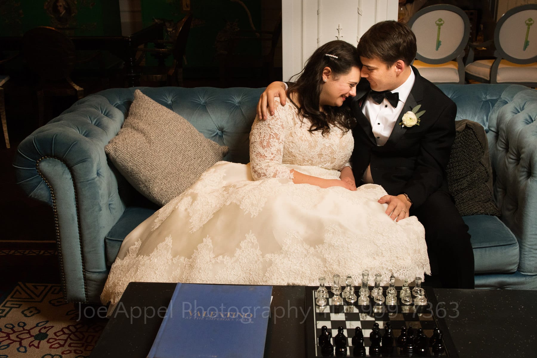 a bride and groom embrace on a blue couch with a chess set and a blue covered book on the table in front of them during their hotel monaco pittsburgh wedding.