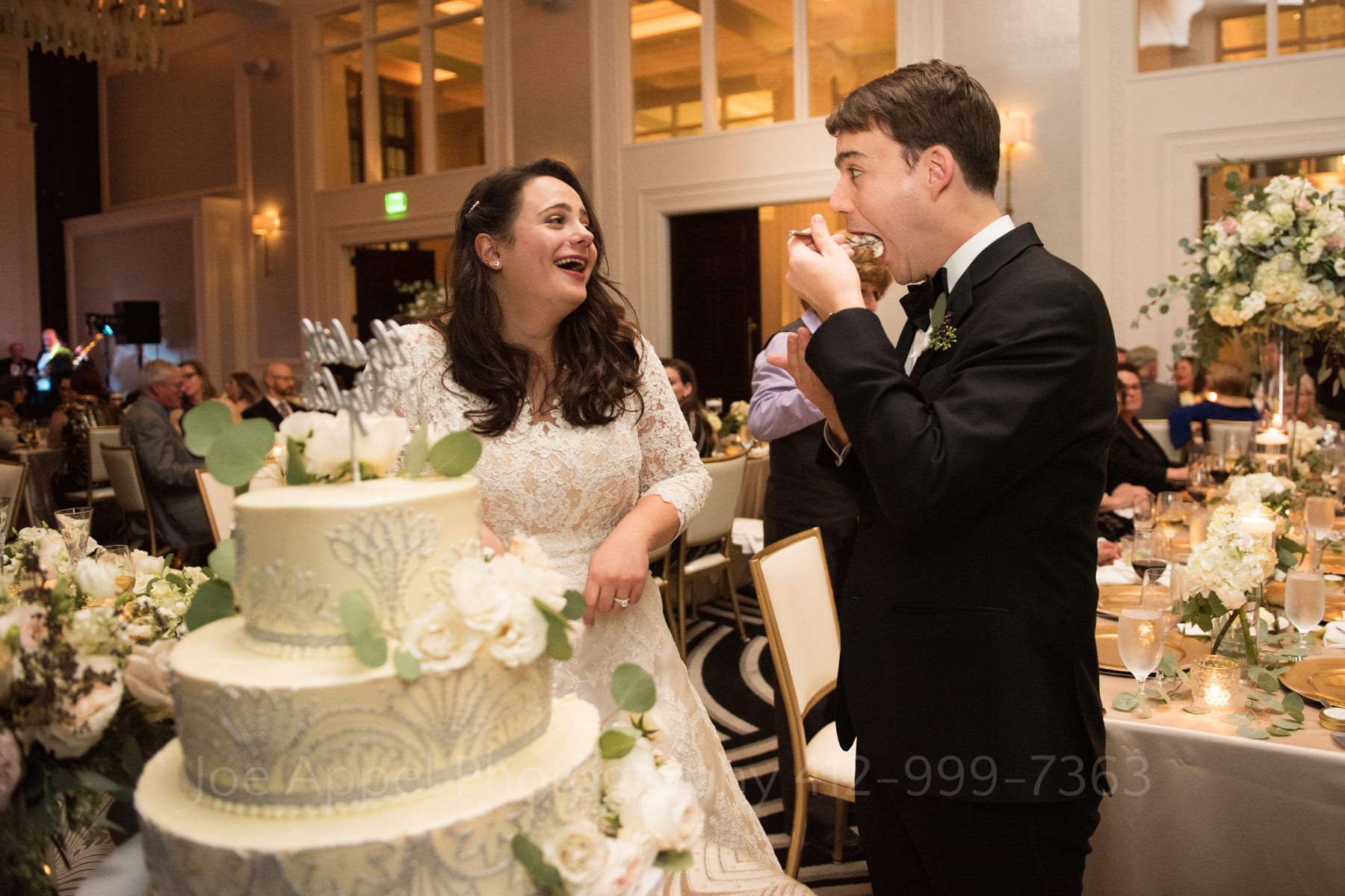 a bride laughs as the groom shoves a huge forkful of their wedding cake into his mouth