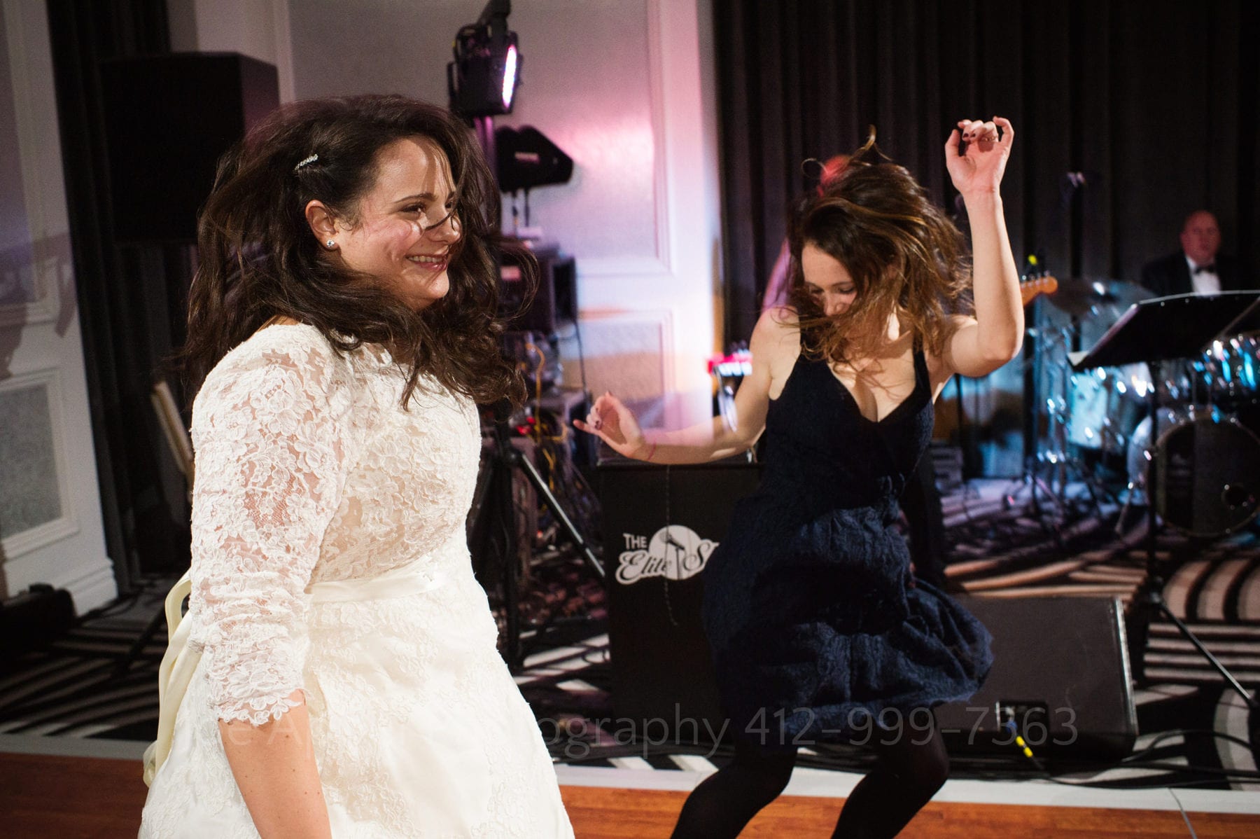 a bride in a white lace dress with sleeves and her friend in a black strap dress jump and dance as their hair flies around them.