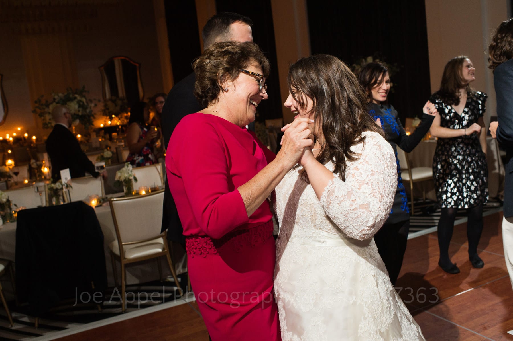 a bride in a white lace dress dances with her mother who is wearing glasses and a red sleeved dress.