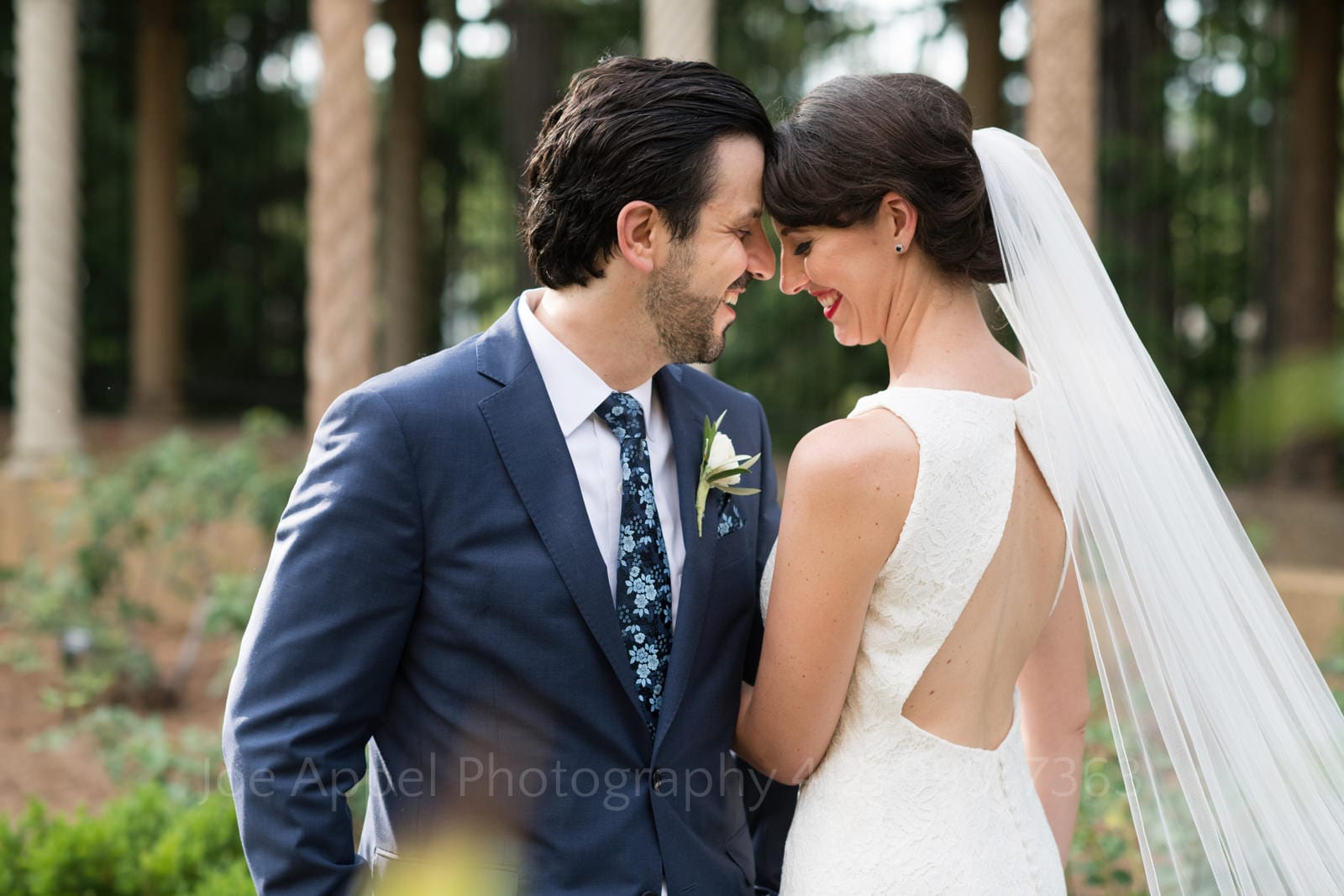 A dark haired groom in a blue suit and blue floral tie smiles has he and his bride with a long veil and open-backed white gown stand with their foreheads touching at their St Francis Hall Washington DC wedding